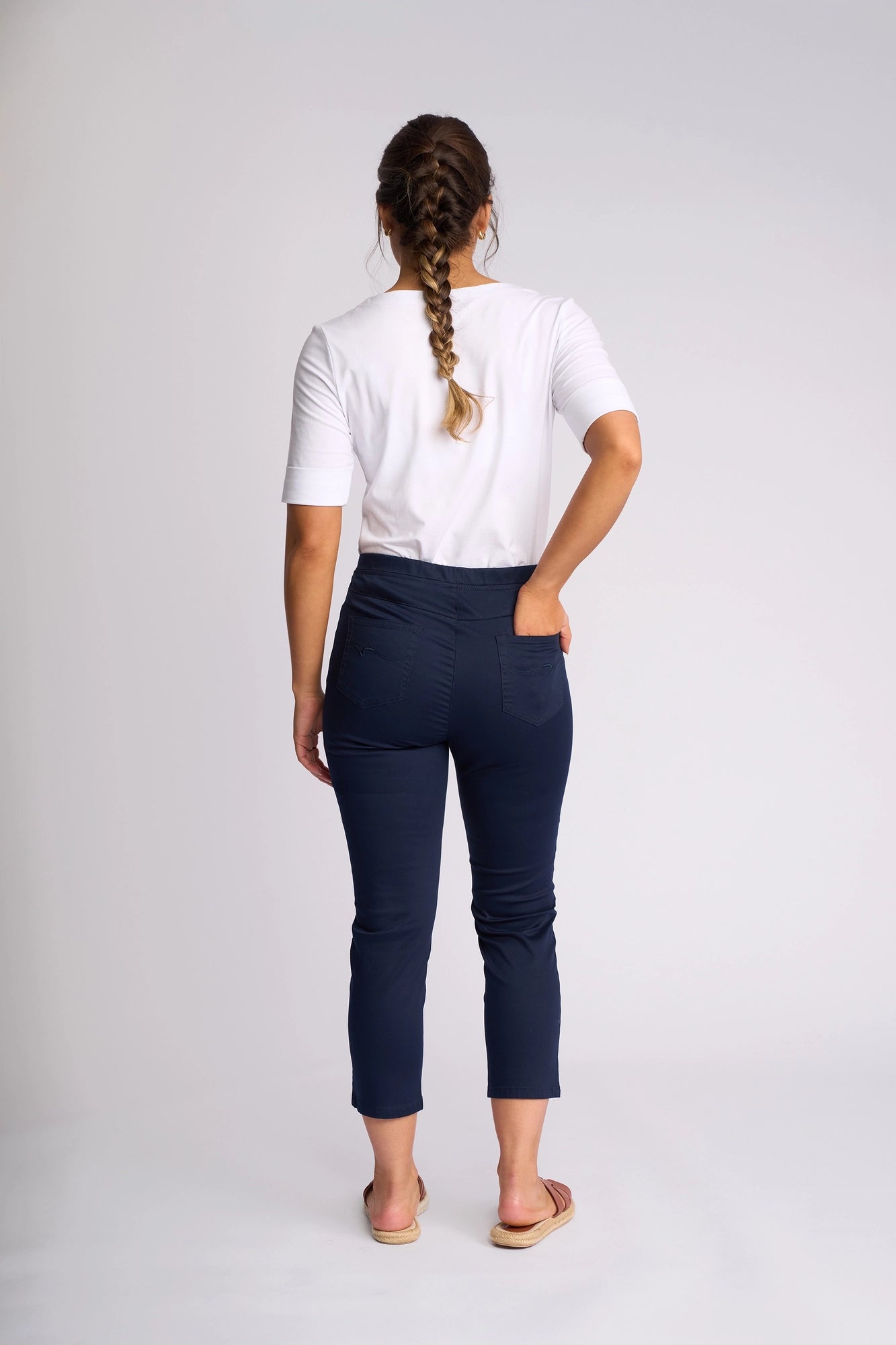 A woman with braided hair stands facing away, wearing a white short-sleeve shirt, Vassalli Harbour 7/8 Pant in navy crafted from stretch fabric, and sandals, against a plain light background.