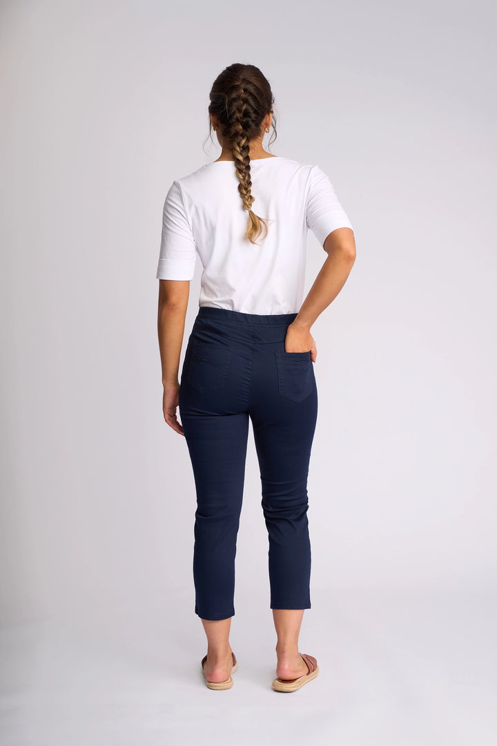 A woman with braided hair stands facing away, wearing a white short-sleeve shirt, Vassalli Harbour 7/8 Pant in navy crafted from stretch fabric, and sandals, against a plain light background.
