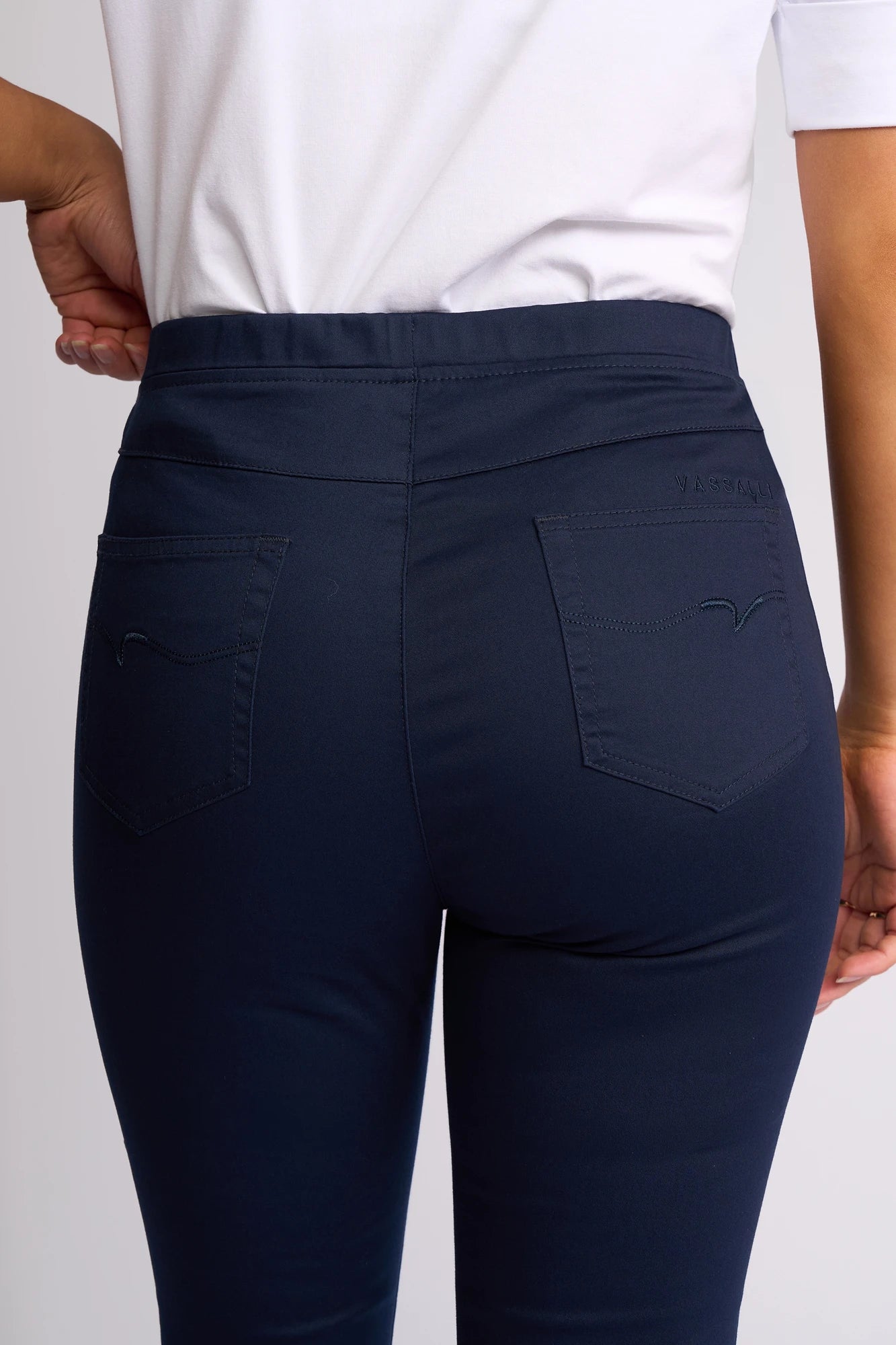 A person is shown from the back wearing a white shirt and Vassalli’s Harbour 7/8 Pant in Navy, with one hand resting on their hip. The fitted pants, crafted from stretch fabric, feature two back pockets and a skinny-leg pull-on design.