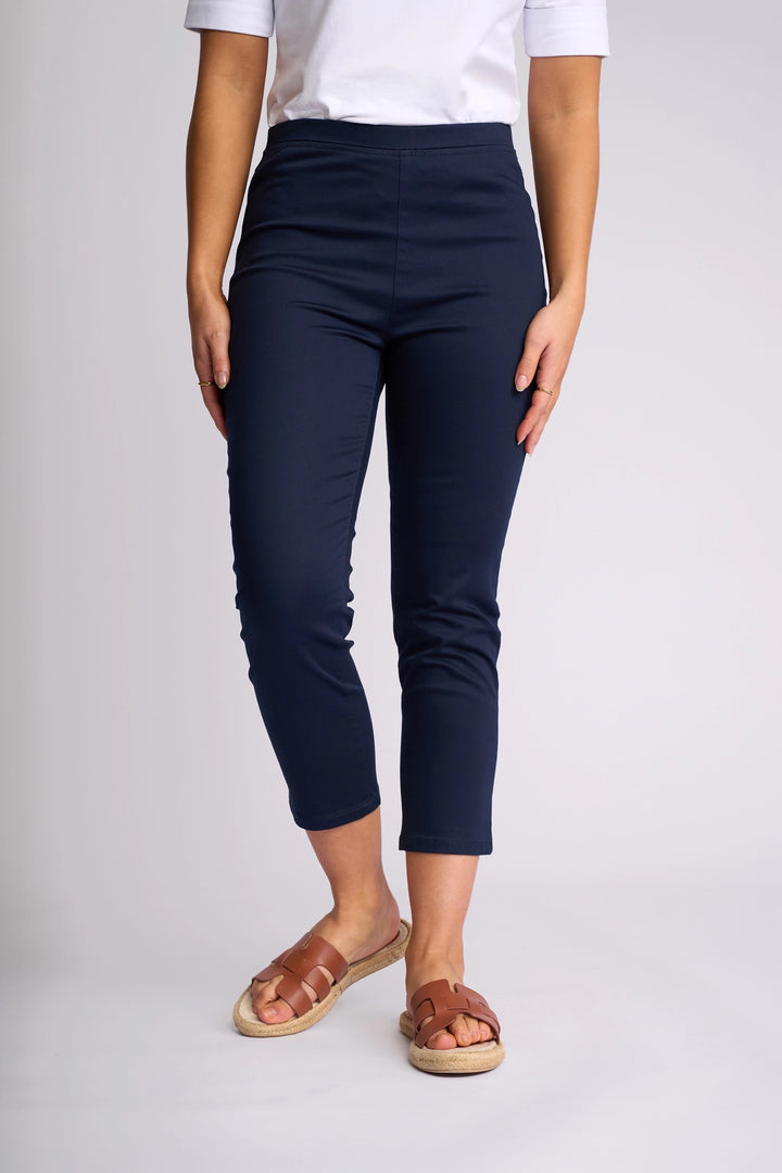 A person wearing a white short-sleeve top, brown sandals, and the Vassalli Harbour 7/8 Pant in navy—crafted from stretch fabric with a skinny-leg, pull-on design—stands against a plain light background.