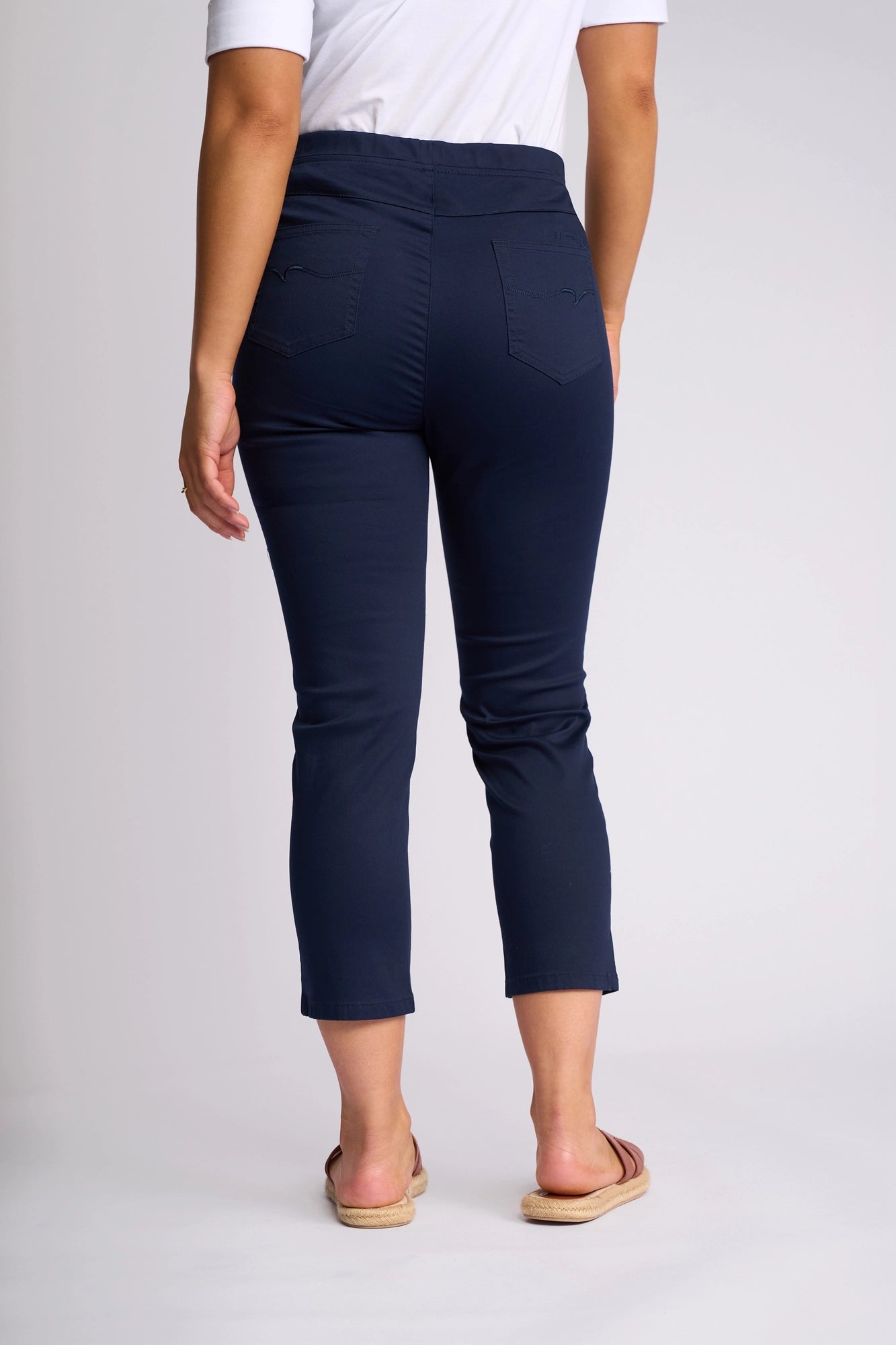 A person wearing a white shirt, Vassalli’s Harbour 7/8 Pant in Navy—skinny-leg pull-on pants crafted from stretch fabric—and sandals is standing with their back to the camera against a plain light background.