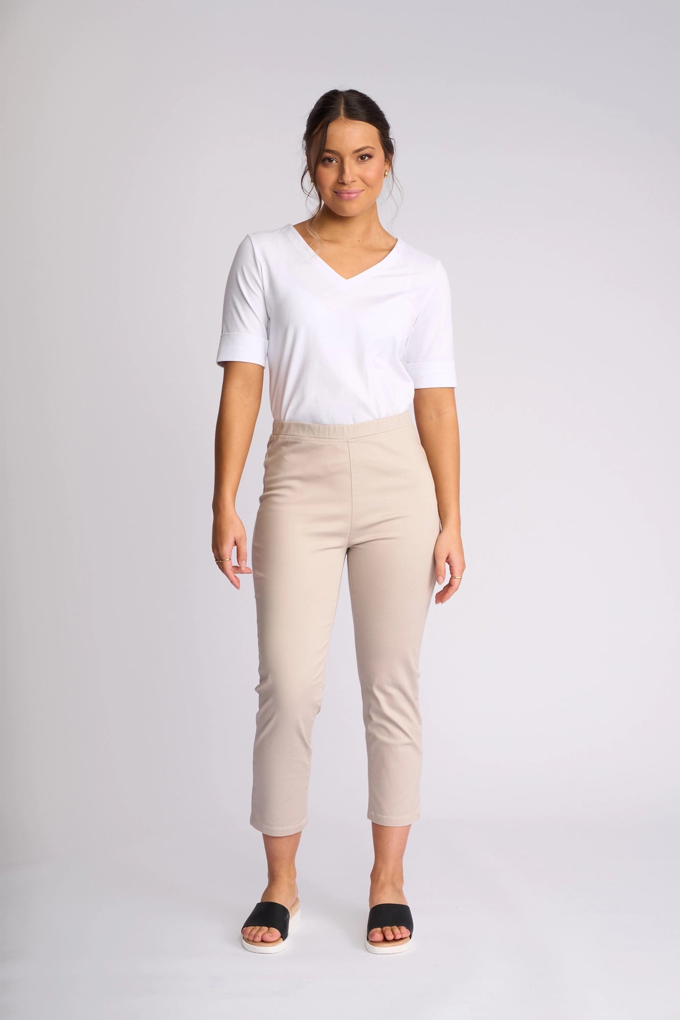 A woman stands facing forward against a plain light background, wearing a white short-sleeve shirt, black slide sandals, and the Vassalli Harbour 7/8 Pant in Pumice.