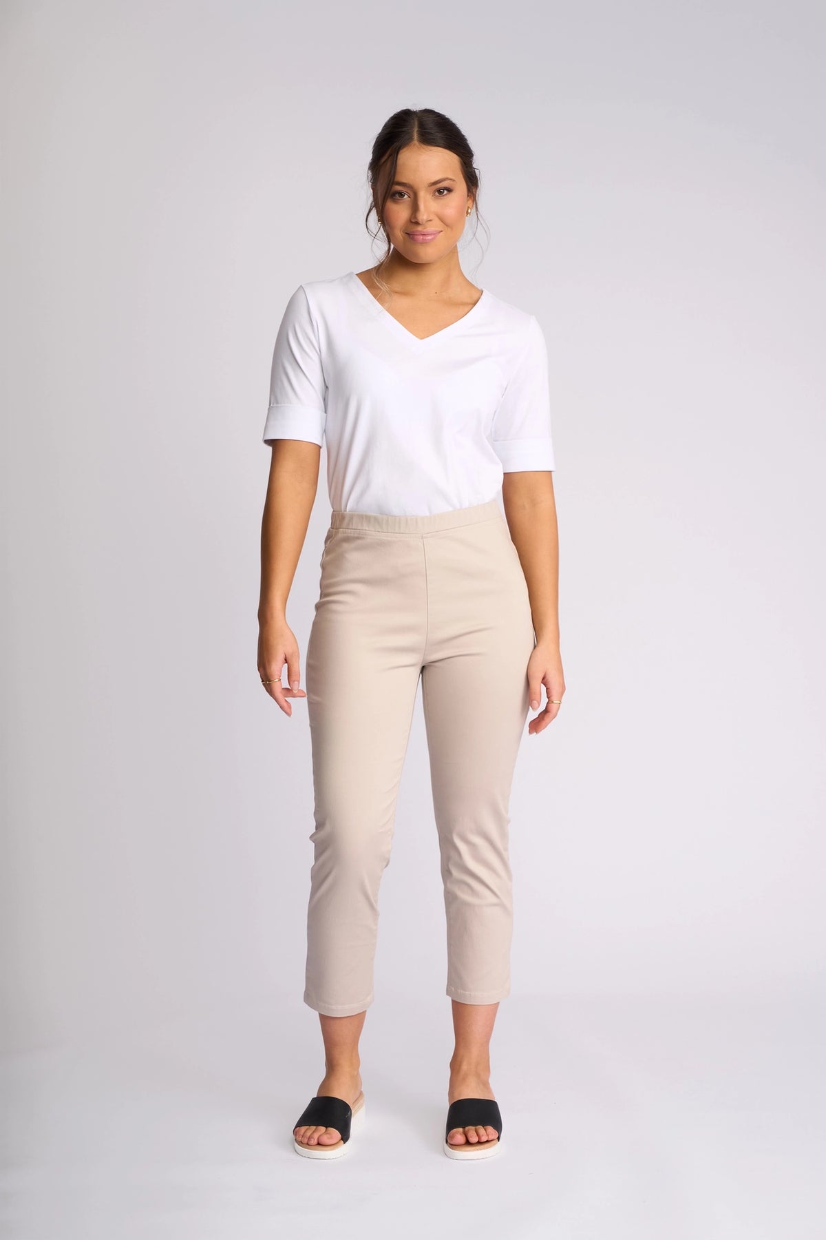 A woman stands facing forward against a plain light background, wearing a white short-sleeve shirt, black slide sandals, and the Vassalli Harbour 7/8 Pant in Pumice.