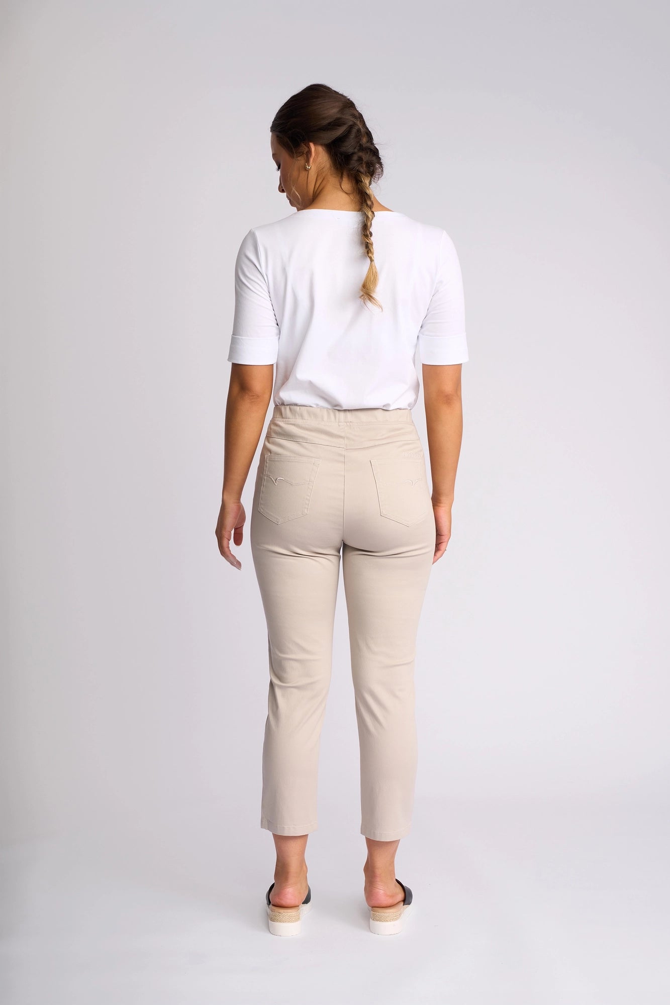A woman stands facing away from the camera in a well-lit studio with a plain background, her hair styled in a braid. She is wearing a white shirt paired with Vassalli’s Harbour 7/8 Pant in Pumice—the chic, beige skinny-leg pull-on pant crafted from stretch fabric for ultimate comfort and style. The 7/8 length pants are elegantly complemented by sandals.