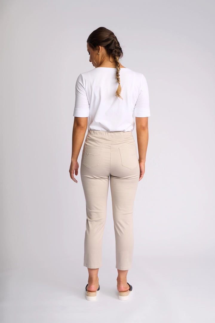 A woman stands facing away from the camera in a well-lit studio with a plain background, her hair styled in a braid. She is wearing a white shirt paired with Vassalli’s Harbour 7/8 Pant in Pumice—the chic, beige skinny-leg pull-on pant crafted from stretch fabric for ultimate comfort and style. The 7/8 length pants are elegantly complemented by sandals.