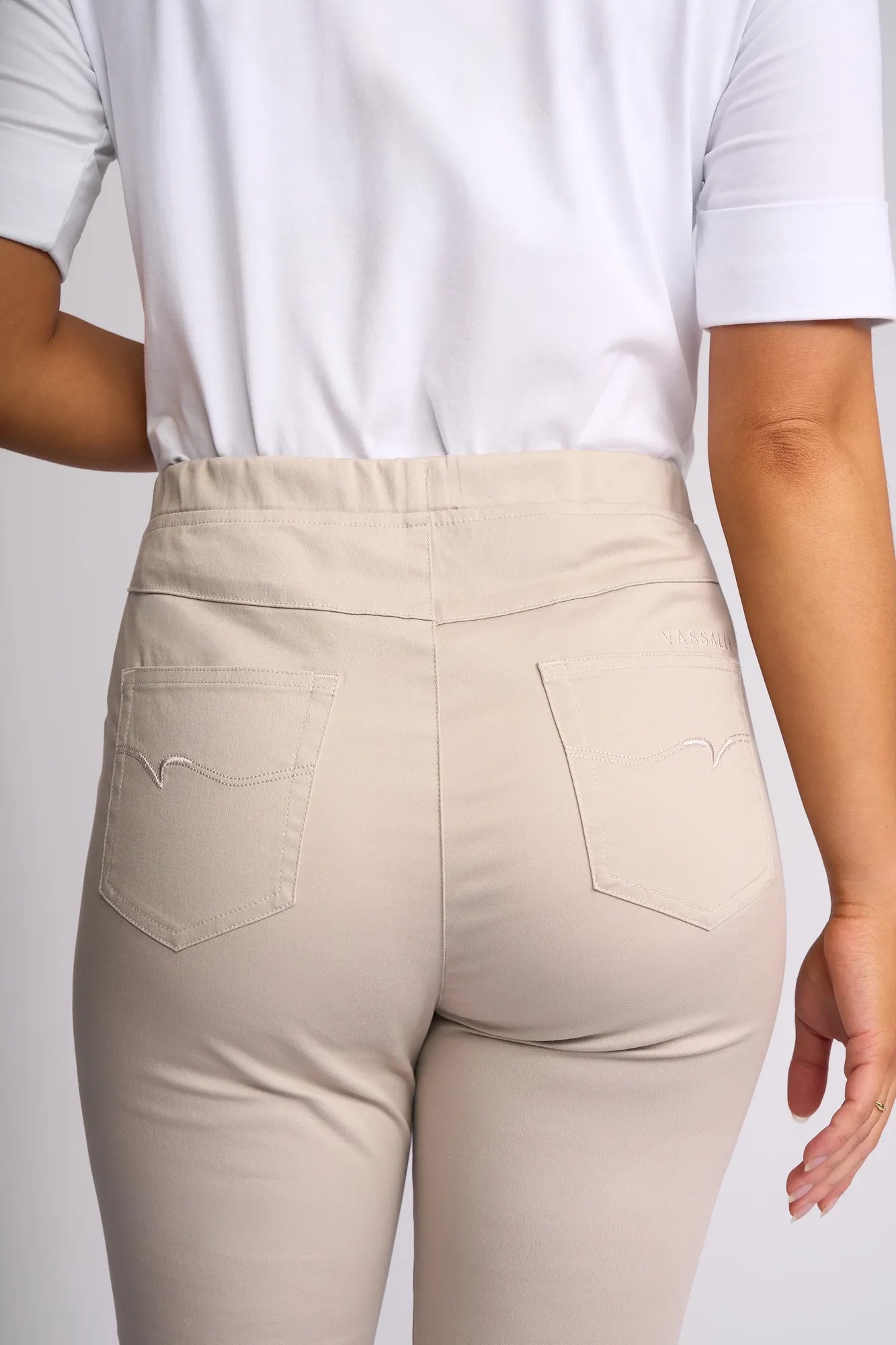 A person is shown from the back wearing the Vassalli Harbour 7/8 Pant in Pumice, featuring a skinny-leg pull-on design crafted from stretch fabric, with two pockets and stitched detailing visible, paired with a white short-sleeve shirt.