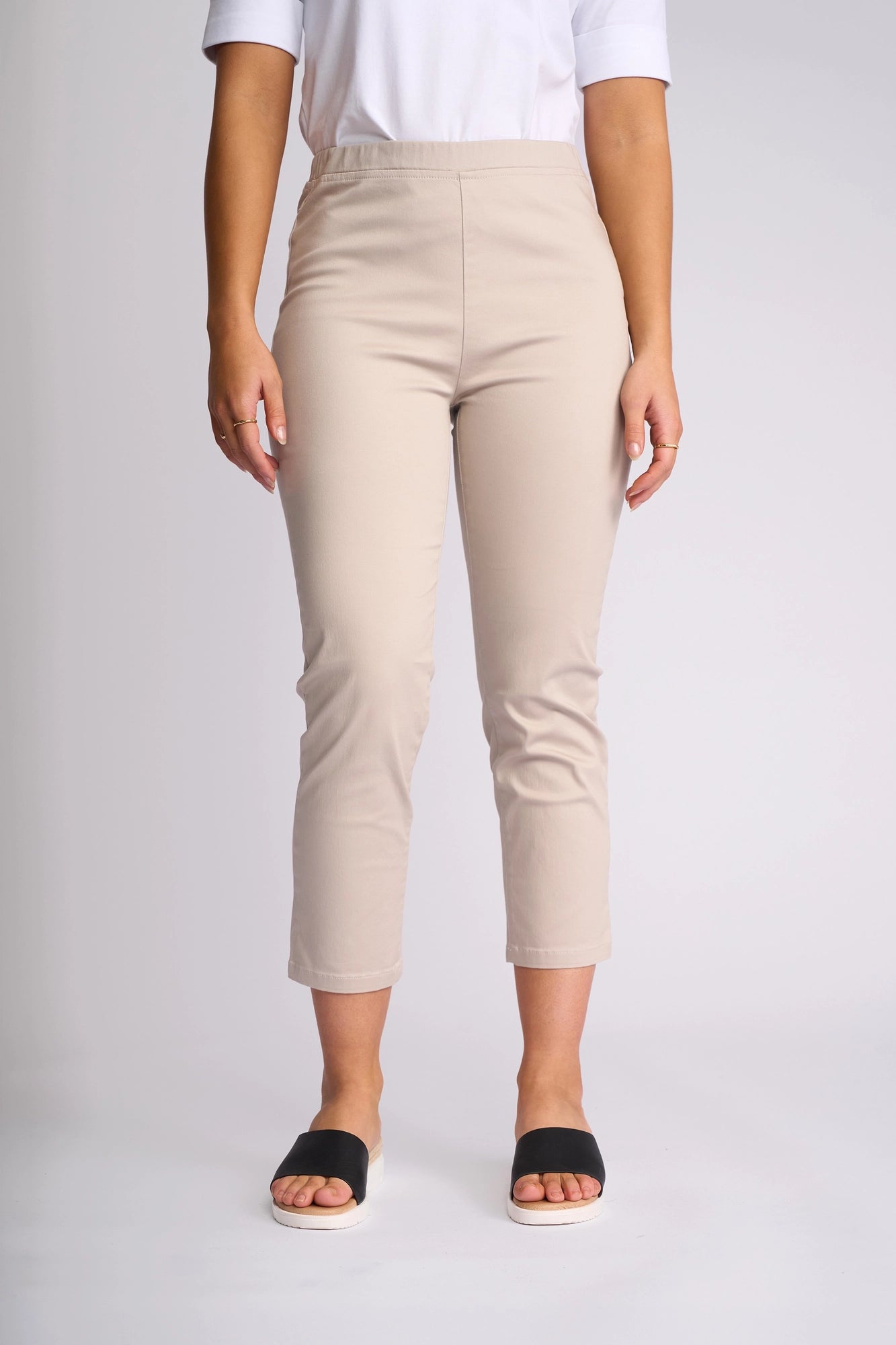 A person is wearing the Vassalli Harbour 7/8 Pant in Pumice, paired with a white shirt and black slide sandals. They are standing against a plain, light background with relaxed arms at their sides, showing only the lower torso and legs.