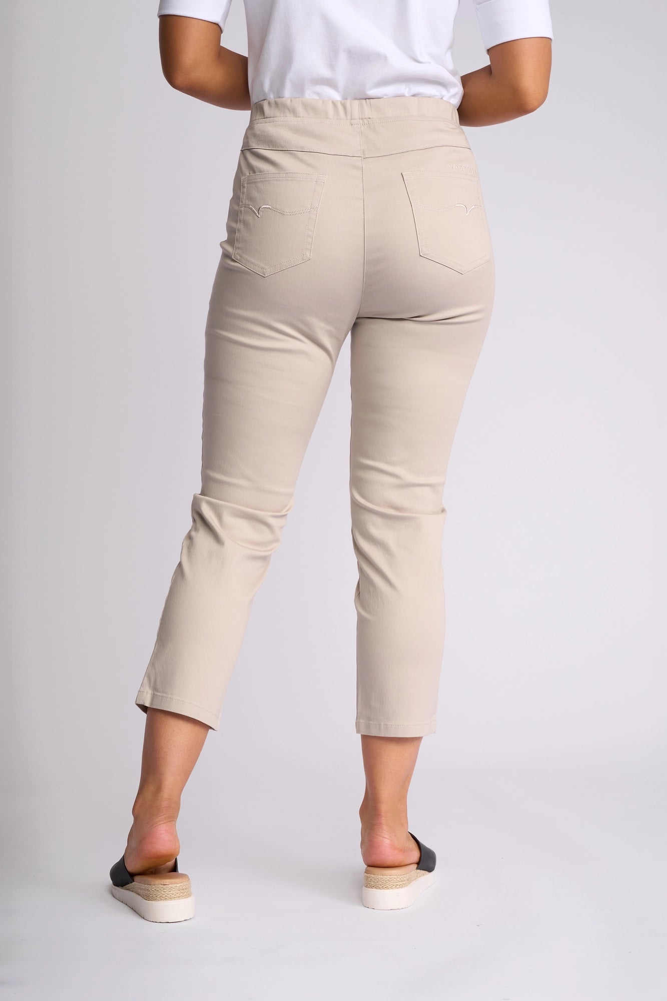A person is seen from behind wearing the Vassalli Harbour 7/8 Pant in Pumice, paired with a white top. The cropped pants, crafted from stretch fabric, feature two back pockets. The outfit is completed with black and white slip-on shoes.