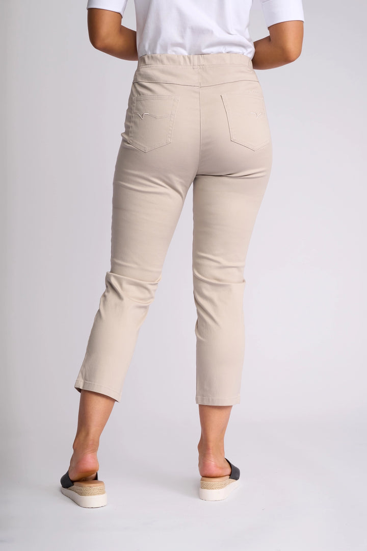 A person is seen from behind wearing the Vassalli Harbour 7/8 Pant in Pumice, paired with a white top. The cropped pants, crafted from stretch fabric, feature two back pockets. The outfit is completed with black and white slip-on shoes.