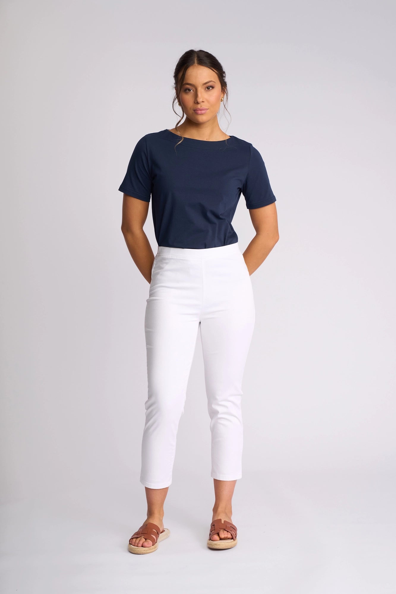 A woman stands against a plain background, wearing the Vassalli Harbour 7/8 Pant in White—skinny-leg pull-on pants crafted from stretch fabric—paired with a navy blue short-sleeve top and brown sandals. She has her hands behind her back and a neutral expression.