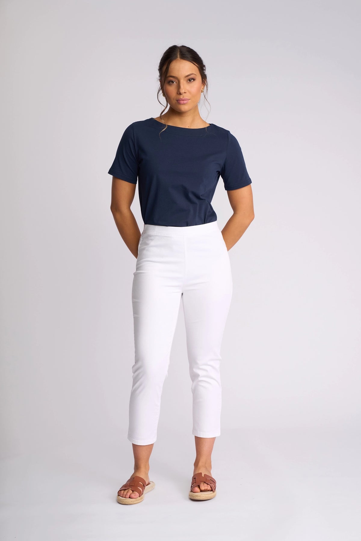 A woman stands against a plain background, wearing the Vassalli Harbour 7/8 Pant in White—skinny-leg pull-on pants crafted from stretch fabric—paired with a navy blue short-sleeve top and brown sandals. She has her hands behind her back and a neutral expression.