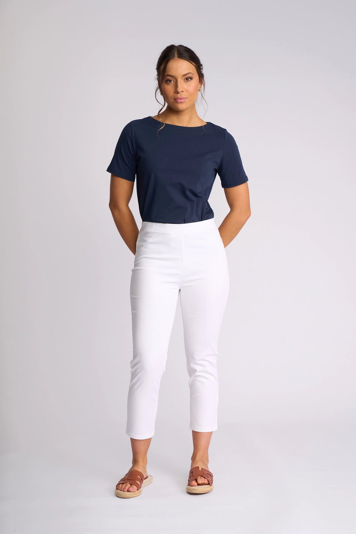 A woman stands against a plain background, wearing the Vassalli Harbour 7/8 Pant in White—skinny-leg pull-on pants crafted from stretch fabric—paired with a navy blue short-sleeve top and brown sandals. She has her hands behind her back and a neutral expression.
