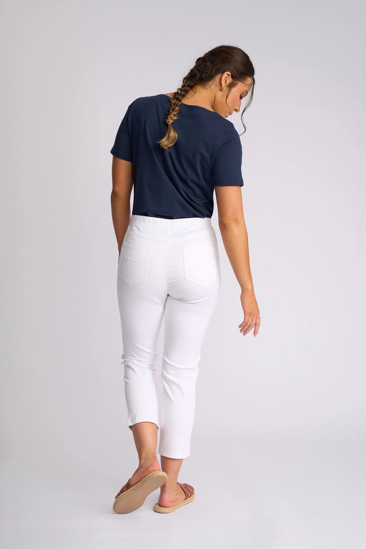 A woman with a braid, wearing a navy t-shirt and Vassalli Harbour 7/8 Pant in white—crafted from stretch fabric in a skinny-leg, pull-on style—pairs the look with sandals as she stands against a plain light background, turned away and looking down over her right shoulder.