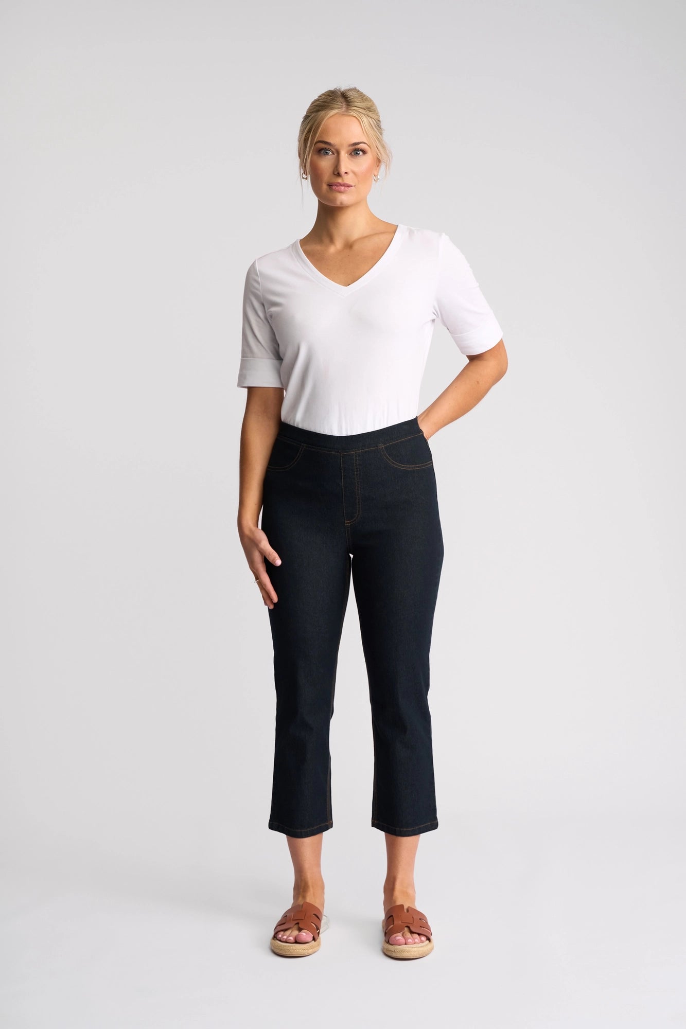 A woman stands against a plain background, wearing a white short-sleeve V-neck top, Vassalli’s Sierra 7/8 Pant in Indigo Denim, and brown sandals. Her right hand is by her side while her left hand rests in her pocket.