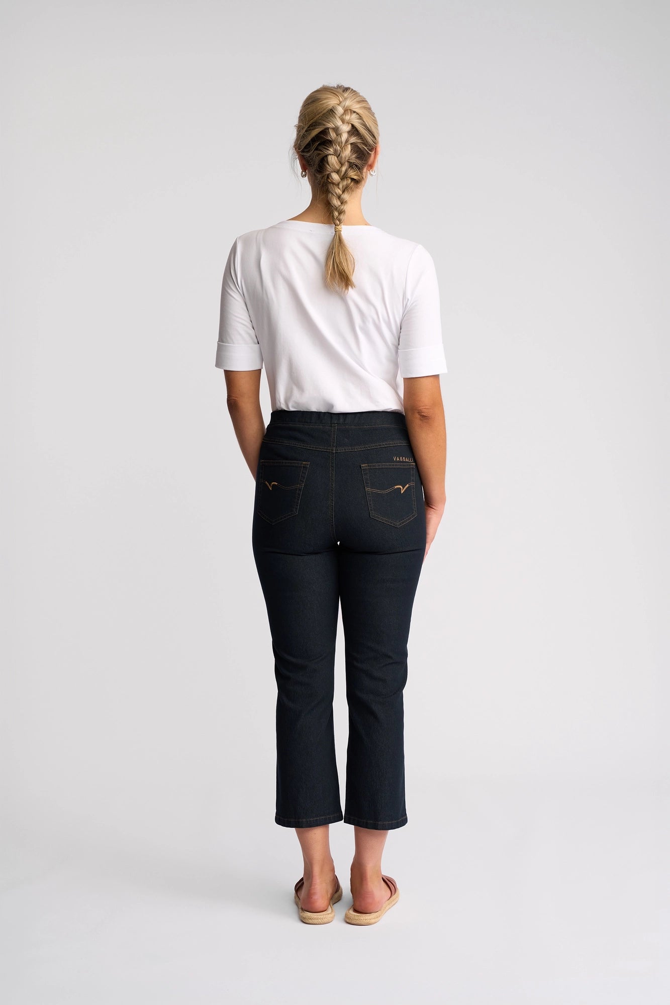 A woman with a blond braid stands facing away, wearing a white shirt and the Vassalli Sierra 7/8 Pant in Indigo Denim, paired with sandals against a plain white background.