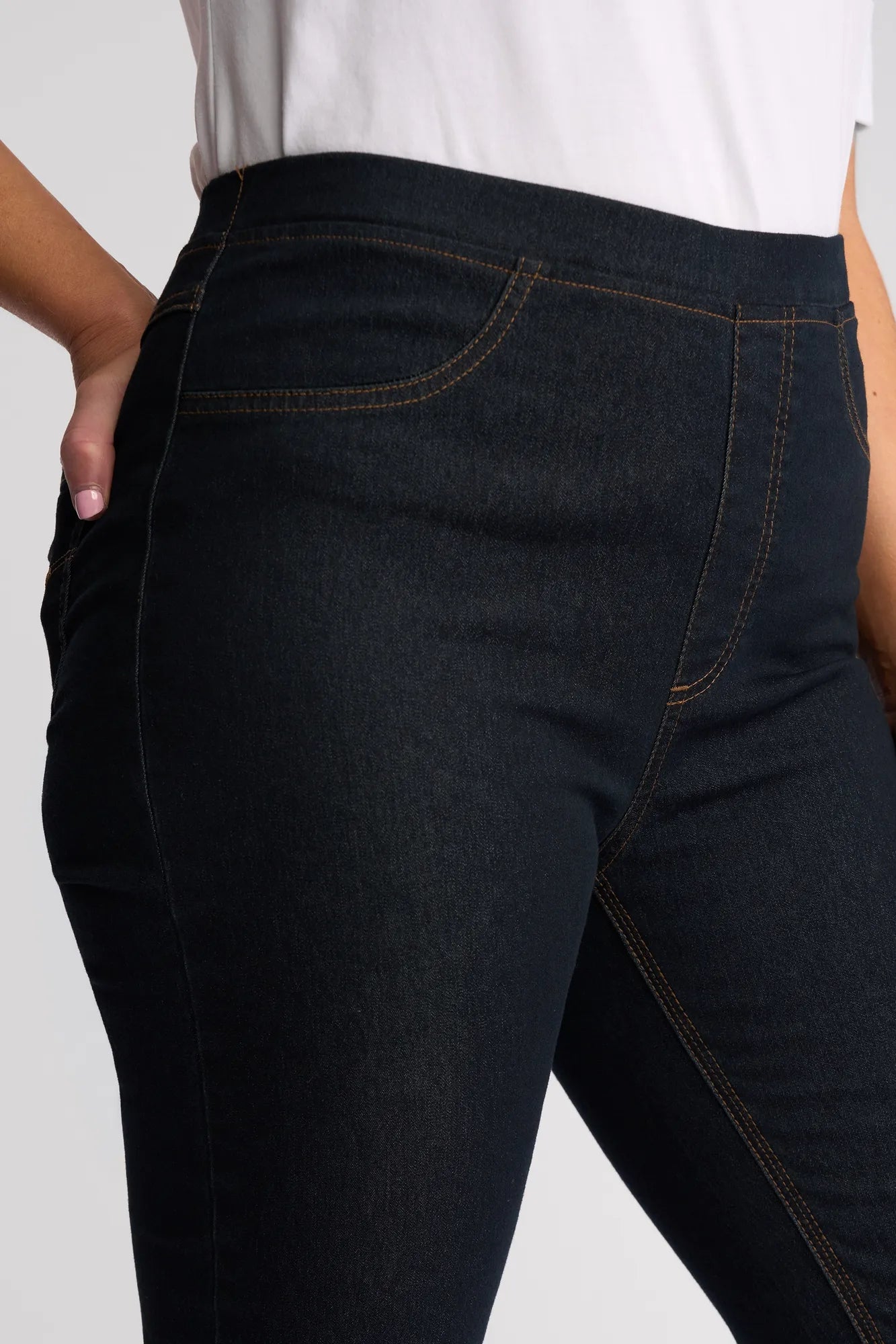 A person wears the Vassalli Sierra 7/8 Pant in Indigo Denim, paired with a white top, with one hand resting on their hip. The image highlights the waist and upper leg area, showcasing the slim-leg silhouette along with detailed stitching and pocket accents of the jeans.
