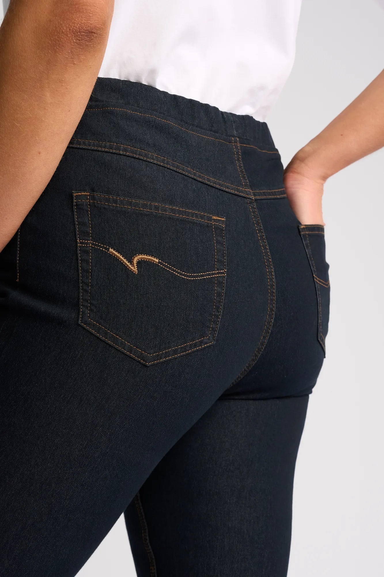 Close-up of a person wearing the Vassalli Sierra 7/8 Pant in indigo denim with yellow stitching and a white shirt, one hand slipped into the back pocket.