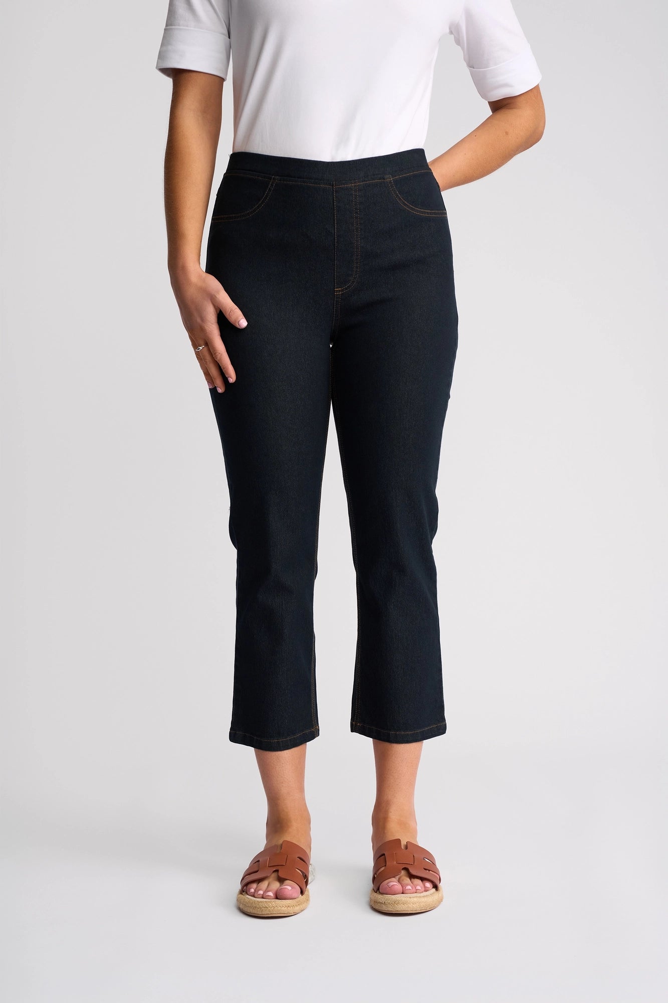 A person wearing a white short-sleeve top, the Vassalli Sierra 7/8 Pant in Indigo Denim, and brown sandals stands with one hand in a pocket against a plain light background.
