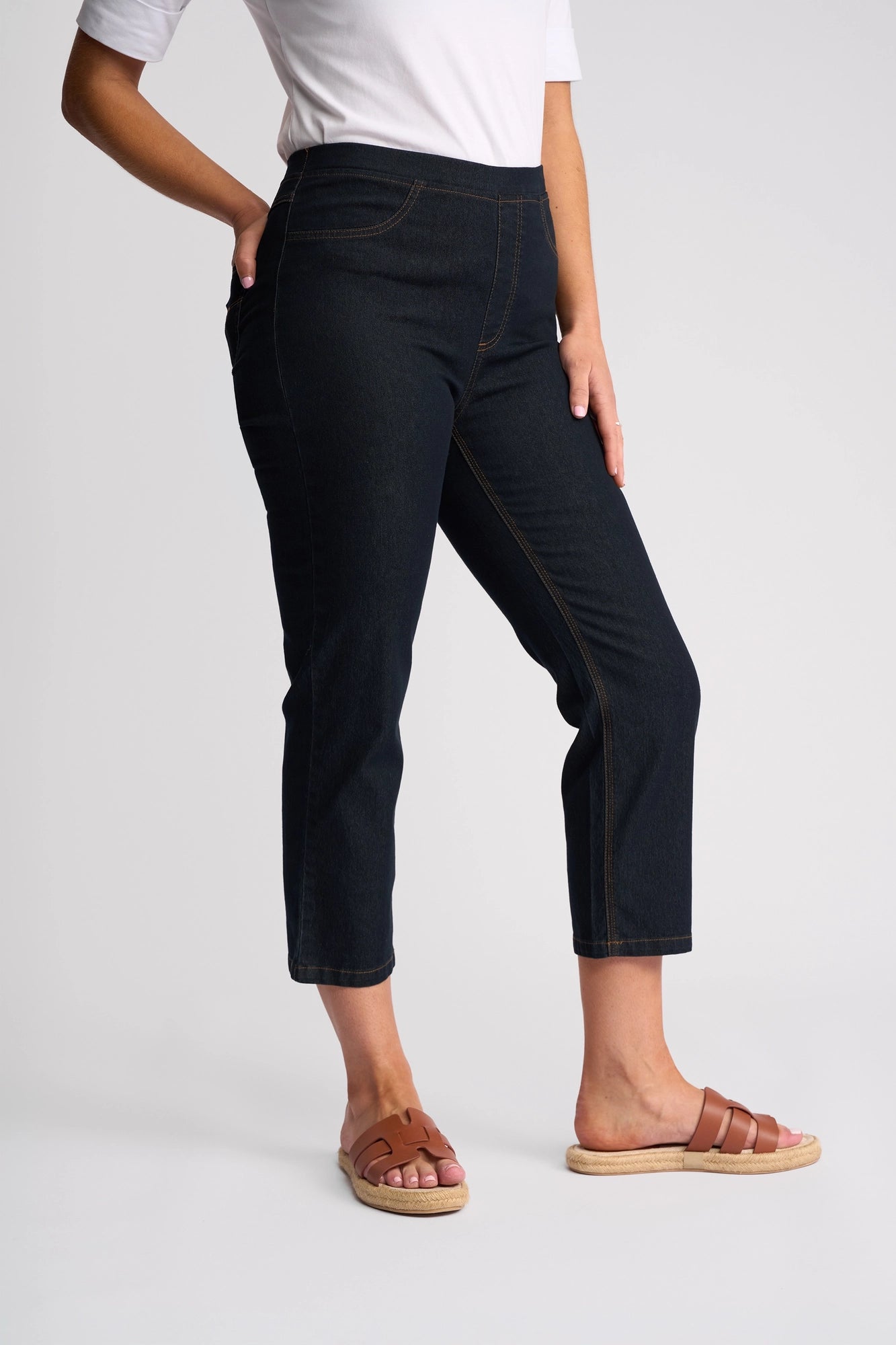 A person wearing a white shirt, Vassalli Sierra 7/8 Pant in Indigo Denim, and tan sandals stands against a plain background with one hand in their pocket.