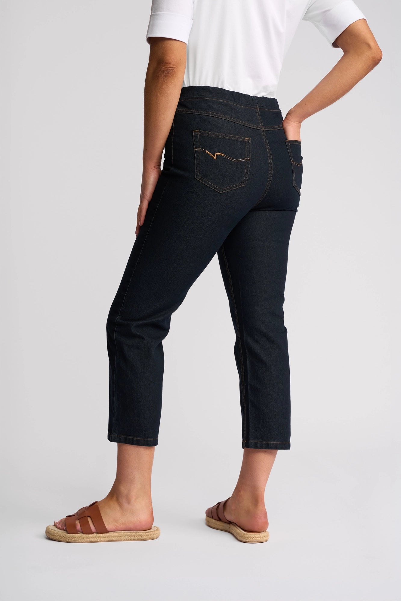 A person wearing a white shirt, Vassalli Sierra 7/8 Pant in Indigo Denim, and brown sandals stands with one hand on their hip, facing away.