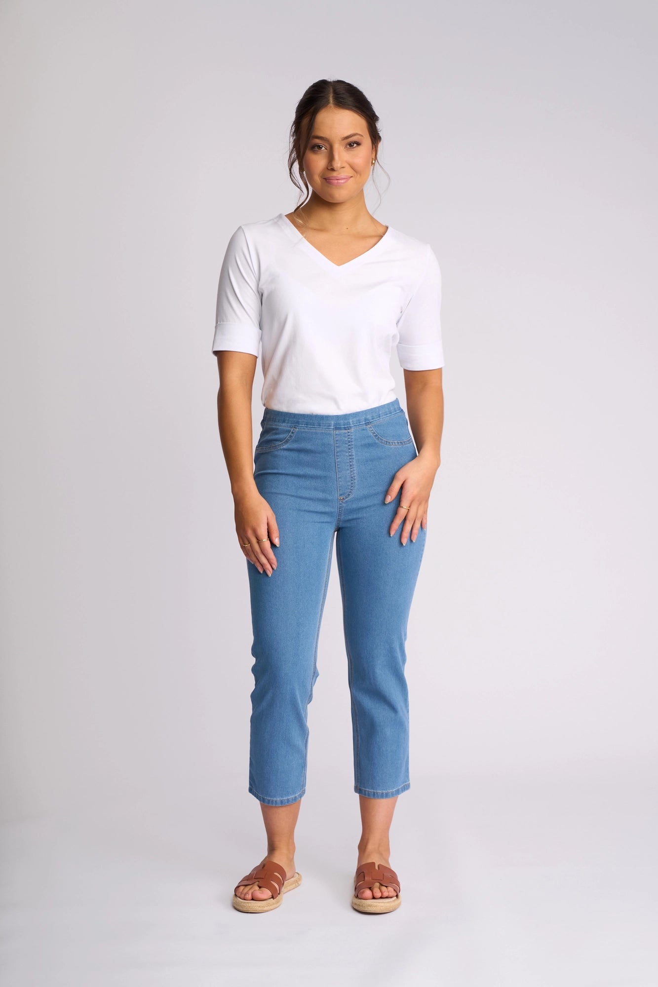A woman stands facing forward, wearing a white V-neck T-shirt, the Vassalli Sierra 7/8 Pant in Light Denim crafted from lightweight stretch denim, and brown sandals. She has dark hair tied back and is posing against a plain light background.