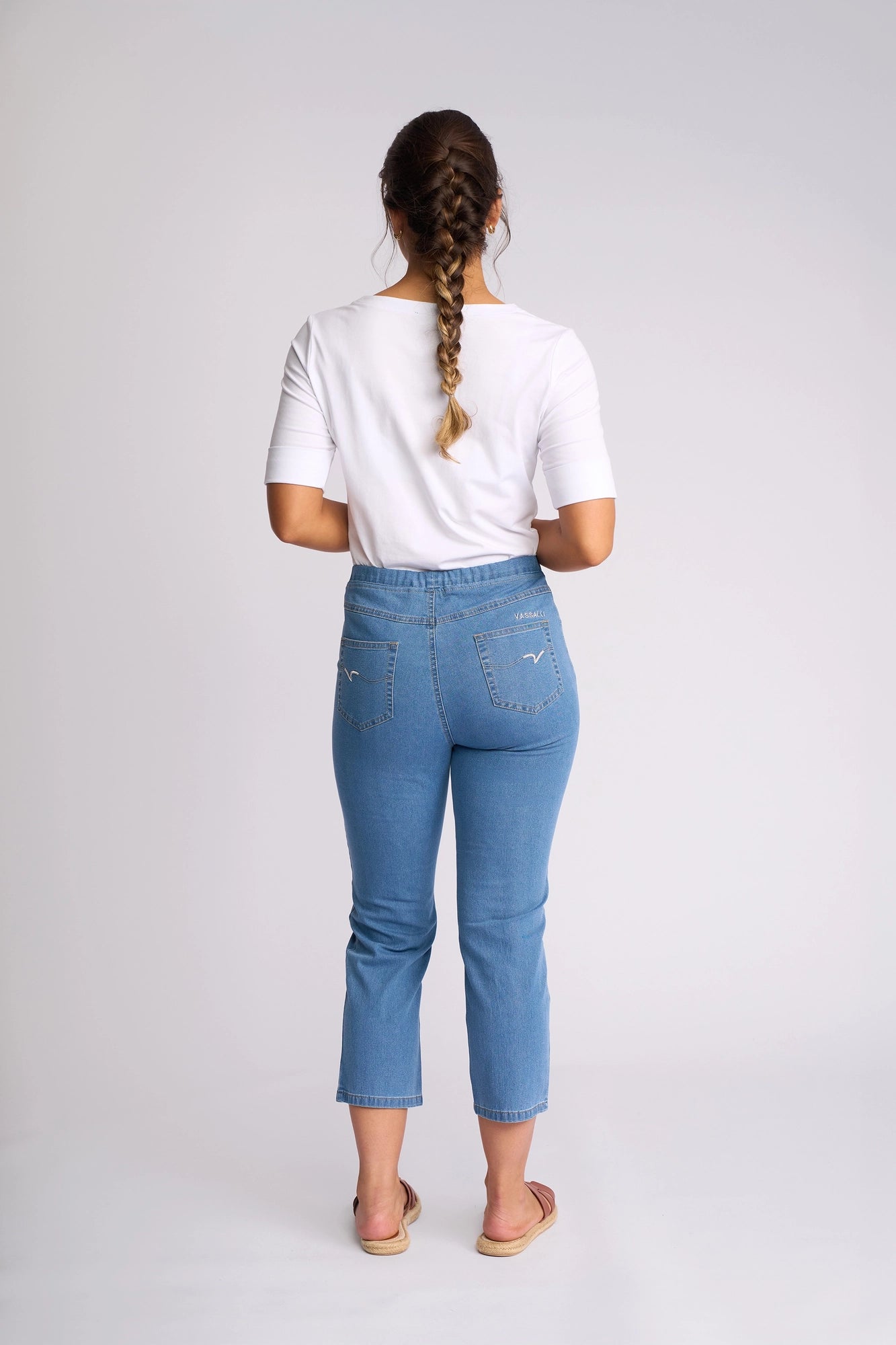 A woman with a braid stands facing away, wearing a white t-shirt, Vassalli Sierra 7/8 Pant in Light Denim made from lightweight stretch denim, and sandals, against a plain white background.