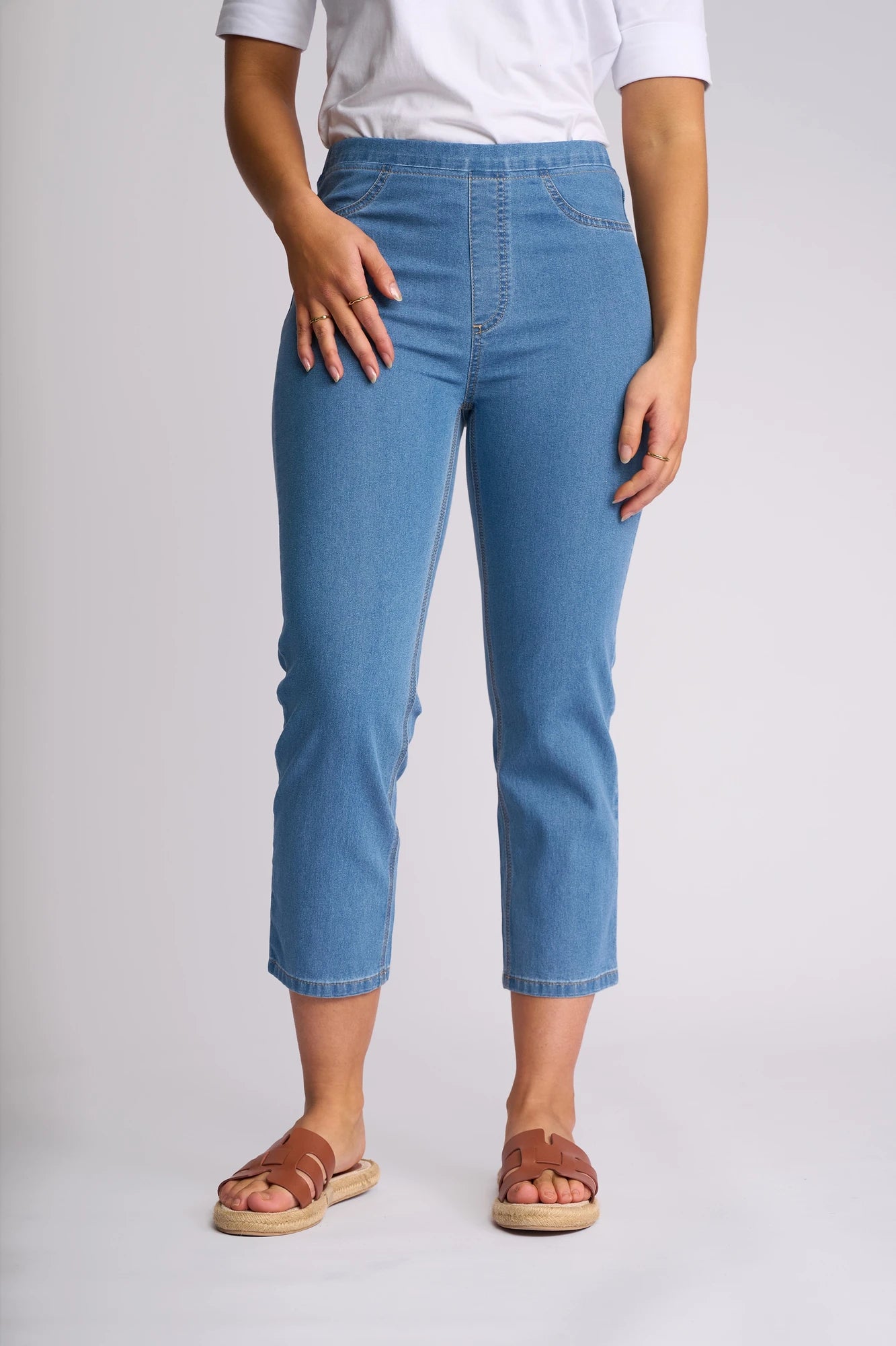 A person wearing a white shirt, Vassalli Sierra 7/8 Pant in Light Denim—crafted from lightweight stretch denim—and brown sandals stands against a plain light background.