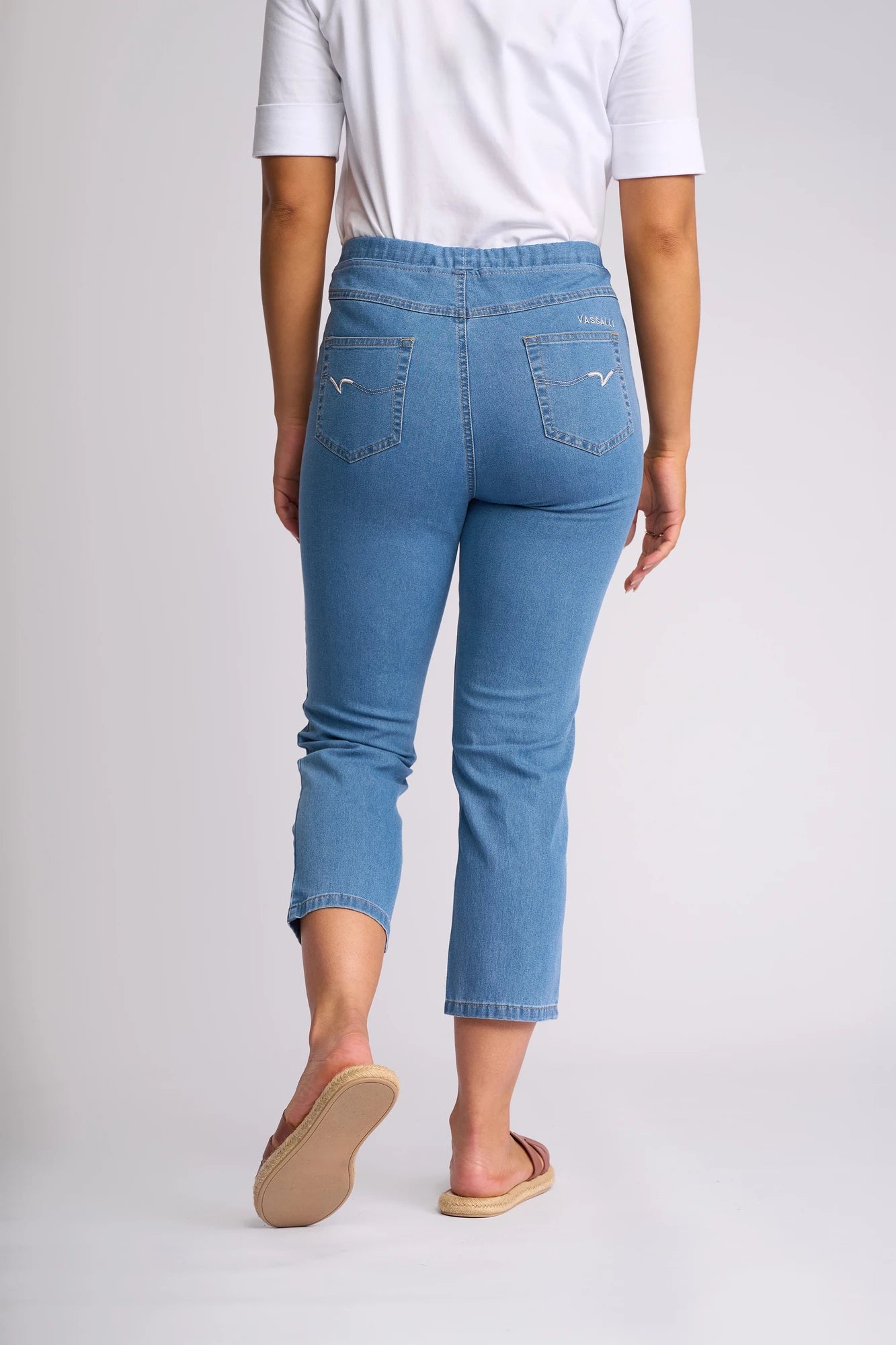 A person stands facing away against a plain light background, wearing a white shirt, sandals, and the Vassalli Sierra 7/8 Pant in Light Denim made from lightweight stretch denim, showcasing the back view of the outfit.