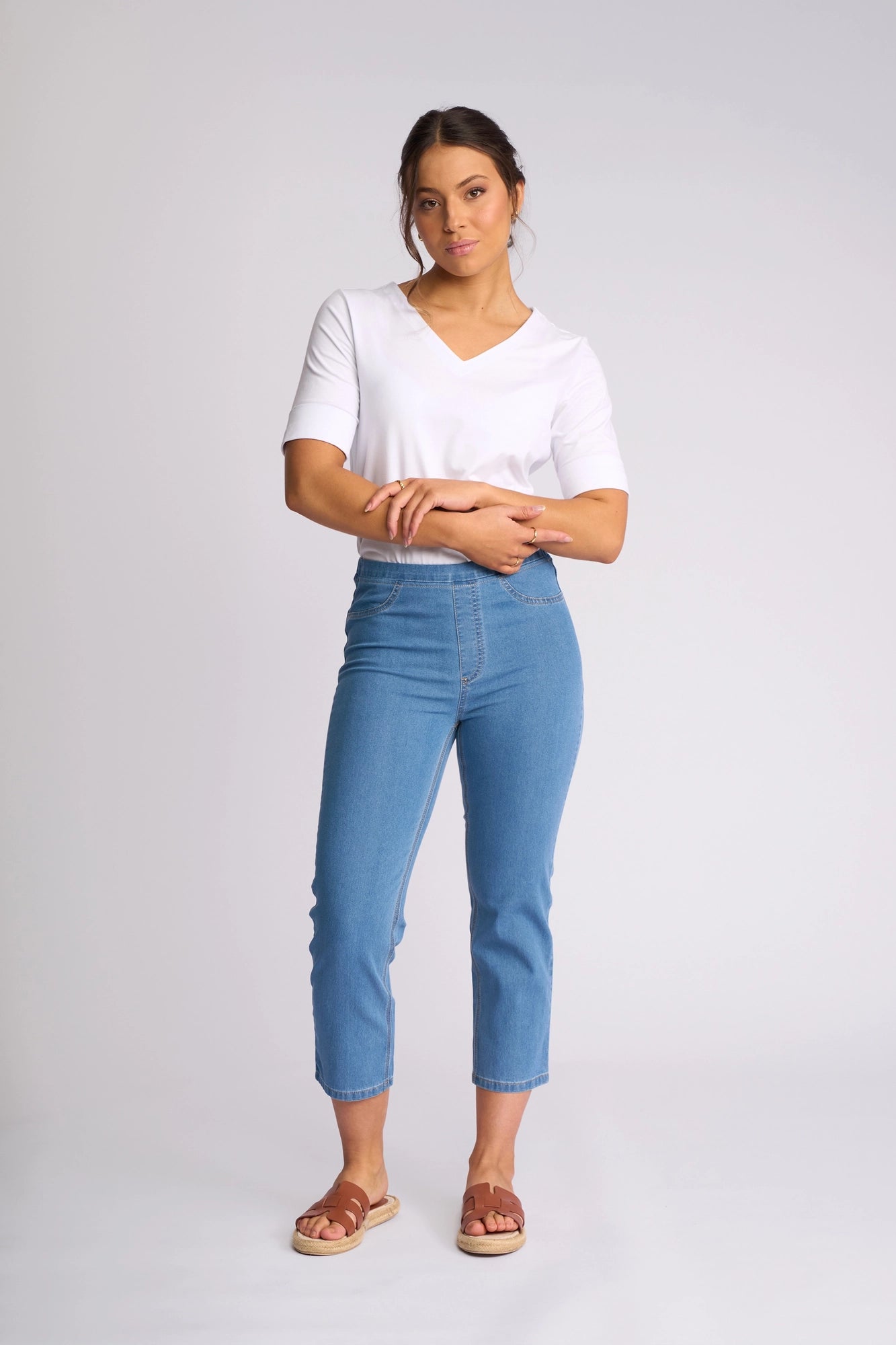 A woman stands against a light background wearing a white short-sleeve top, the Vassalli Sierra 7/8 Pant in Light Denim—high-waisted slim-leg pull-on jeans crafted from lightweight stretch denim—and brown sandals. Her arms are gently crossed as she looks forward with a neutral expression.