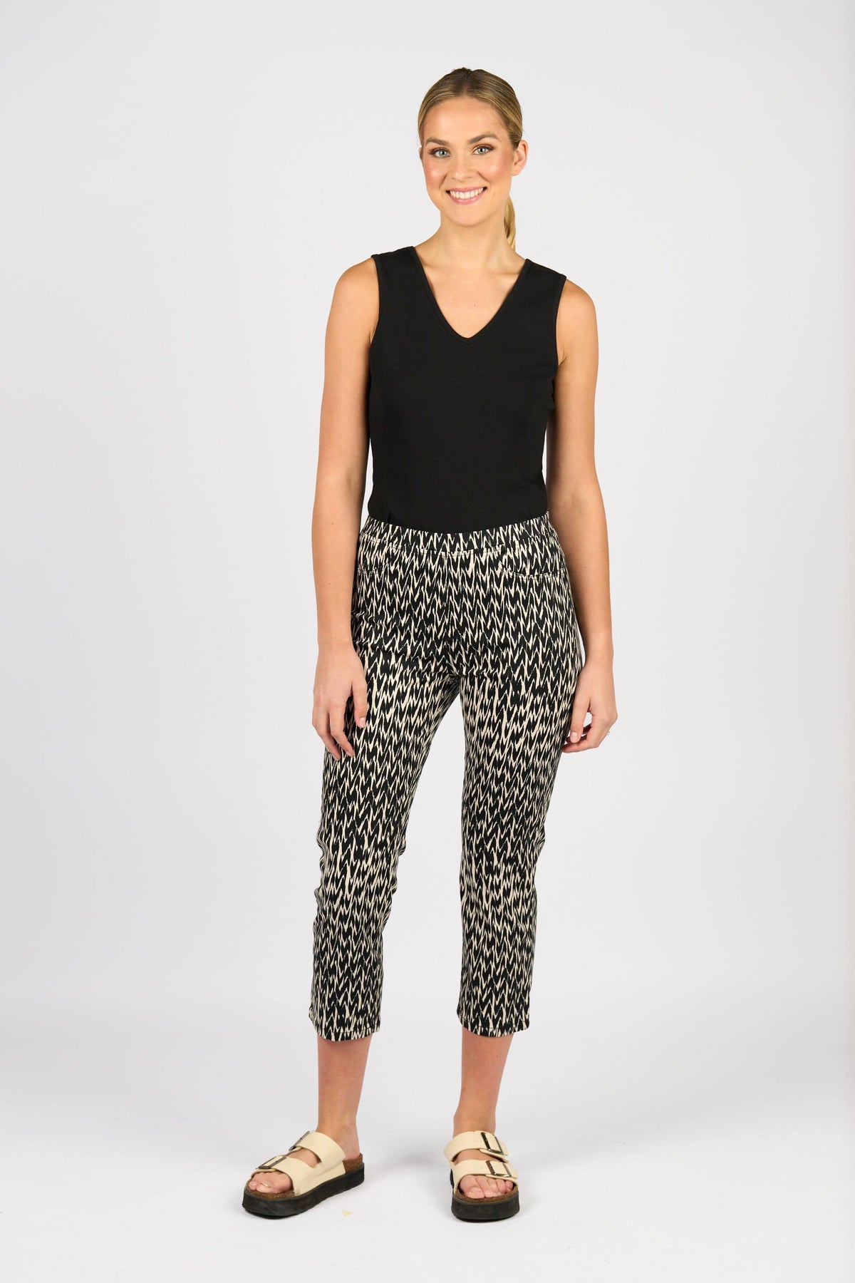 A woman smiles in a sleeveless black top and Vassalli Sierra 7/8 Pant – Beau, paired with beige sandals against a plain light background.