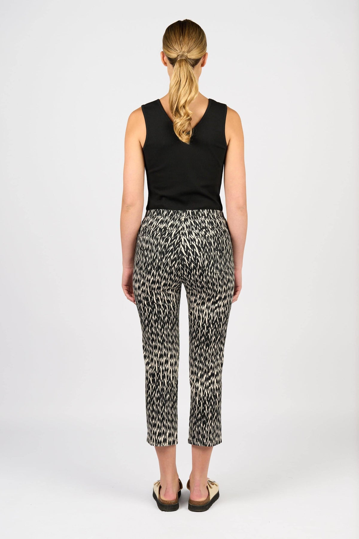 A woman stands with her back to the camera, wearing a black sleeveless top and Vassalli Sierra 7/8 Pant - Beau, styled with black sandals. Her blond hair is in a low ponytail against a plain white background.