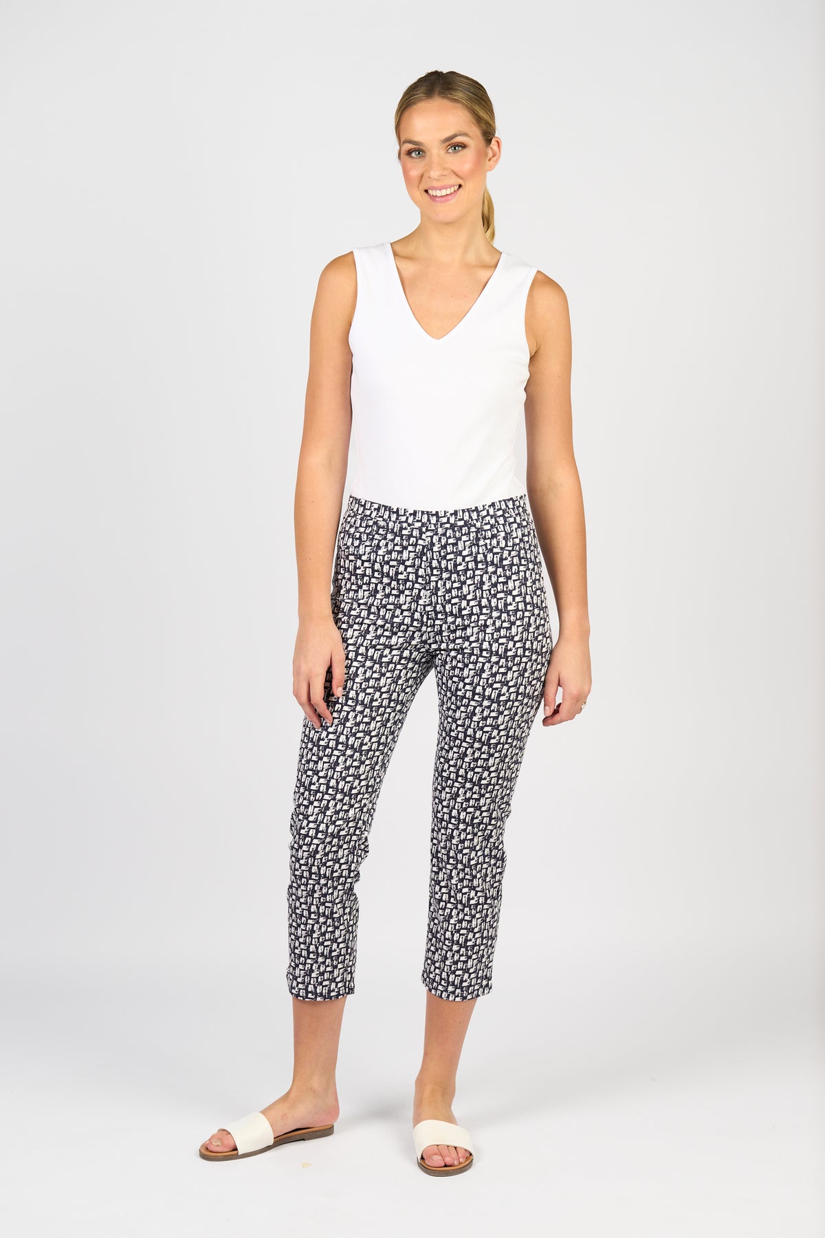 A woman smiles against a plain background, wearing a sleeveless white top, white slide sandals, and Vassalli’s Sierra 7/8 Pant - Cove in a black and white pattern.