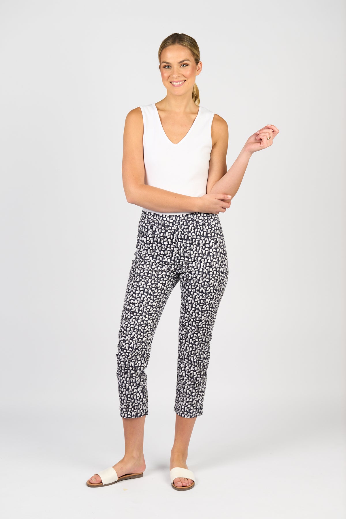 A woman smiles as she poses against a white background, wearing Vassalli's Sierra 7/8 Pant - Cove with a sleeveless white top and white slide sandals. Her hair is styled in a low ponytail.