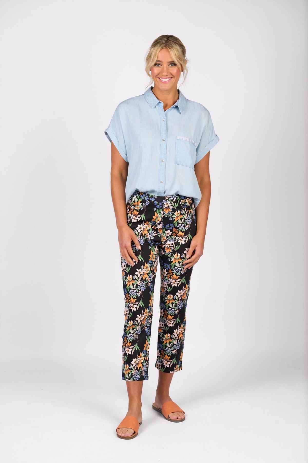 A woman smiles against a plain white background, wearing a light blue short-sleeve button-up shirt, Vassalli Sierra 7/8 Pant - Grove with a floral print, and brown sandals.