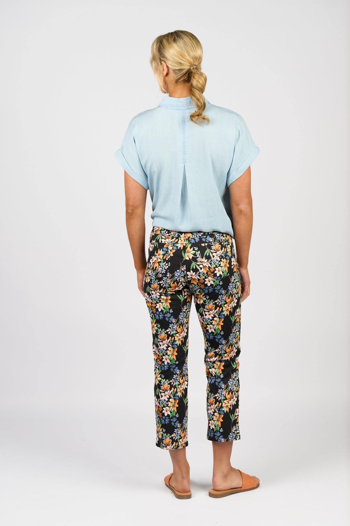 A person stands facing away, wearing a light blue short-sleeve shirt, tan slip-on shoes, and Vassalli Sierra 7/8 Pant - Grove, with blonde hair in a low ponytail against a plain white background.