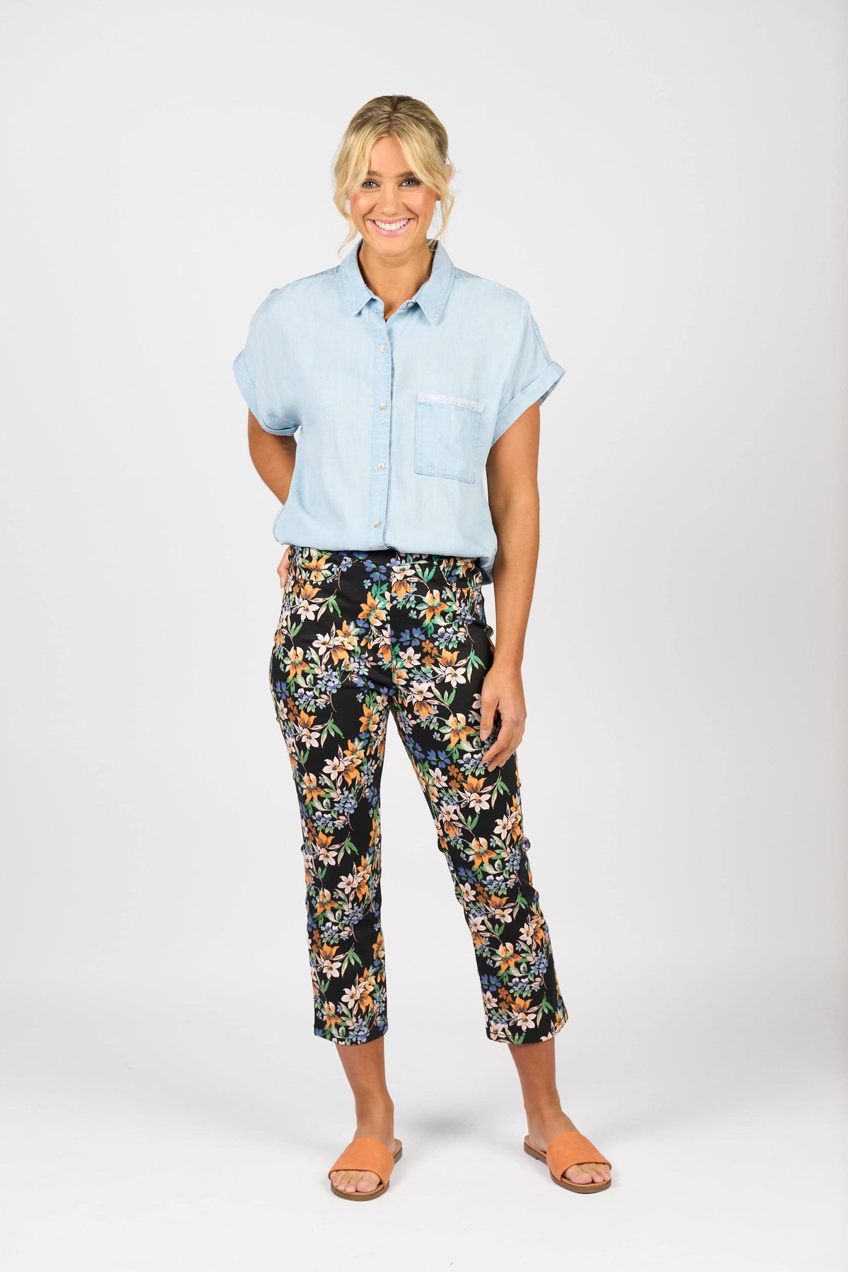 A woman smiles barefoot against a white background, wearing Vassalli's Sierra 7/8 Pant - Grove with a colorful floral print and a light blue short-sleeve button-up shirt.