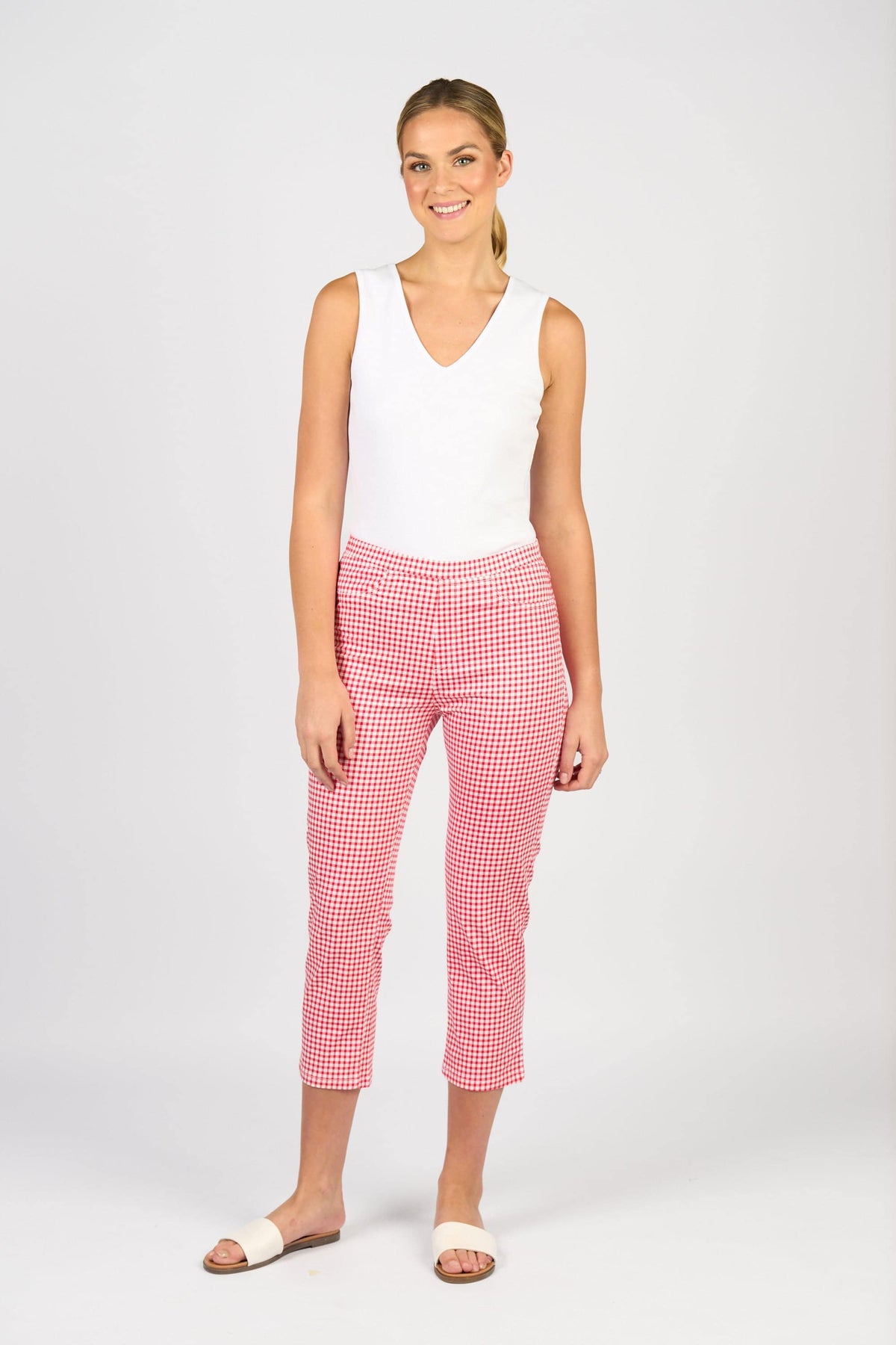 A woman smiles against a plain white background, wearing a sleeveless white V-neck top, white sandals, and Vassalli's Sierra 7/8 Pant in Red Gingham.