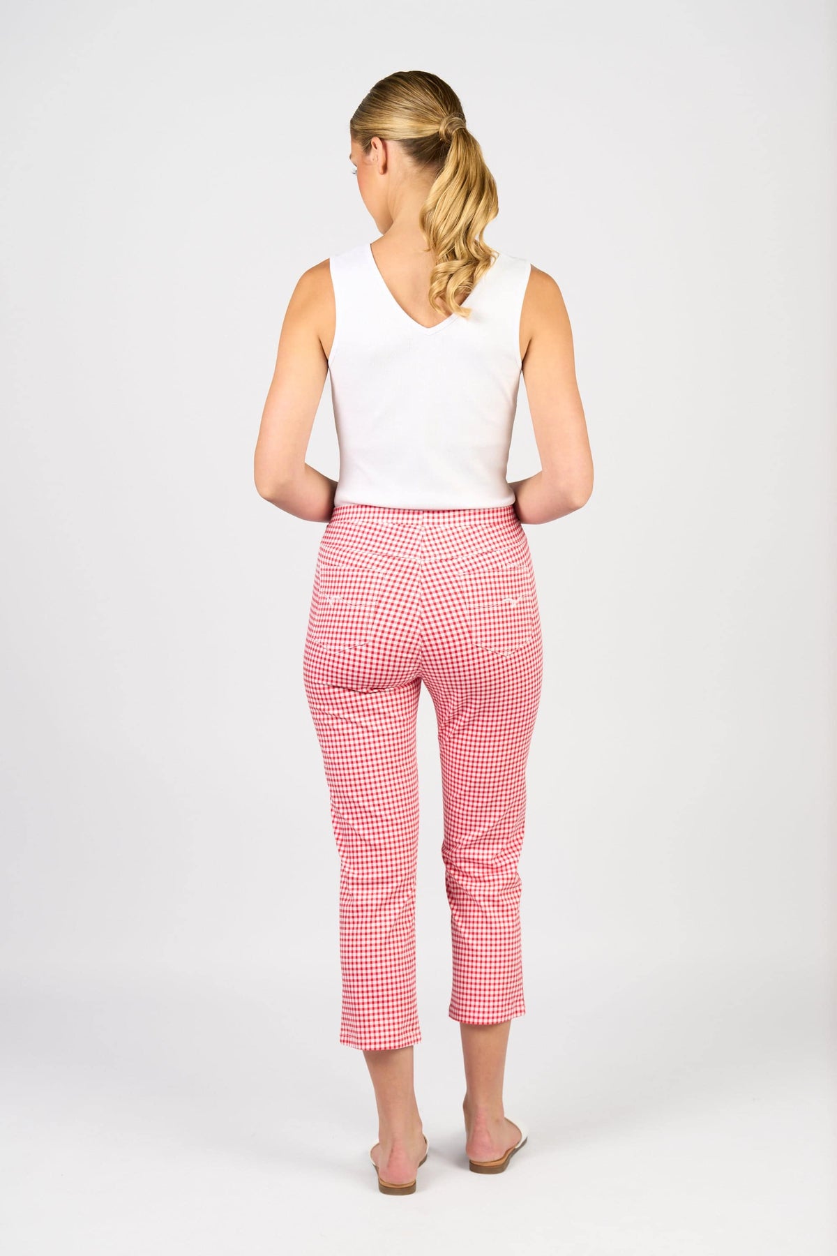 A woman with blonde hair in a ponytail stands barefoot against a plain white background, wearing a white sleeveless top and Vassalli Sierra 7/8 Pant in Red Gingham.