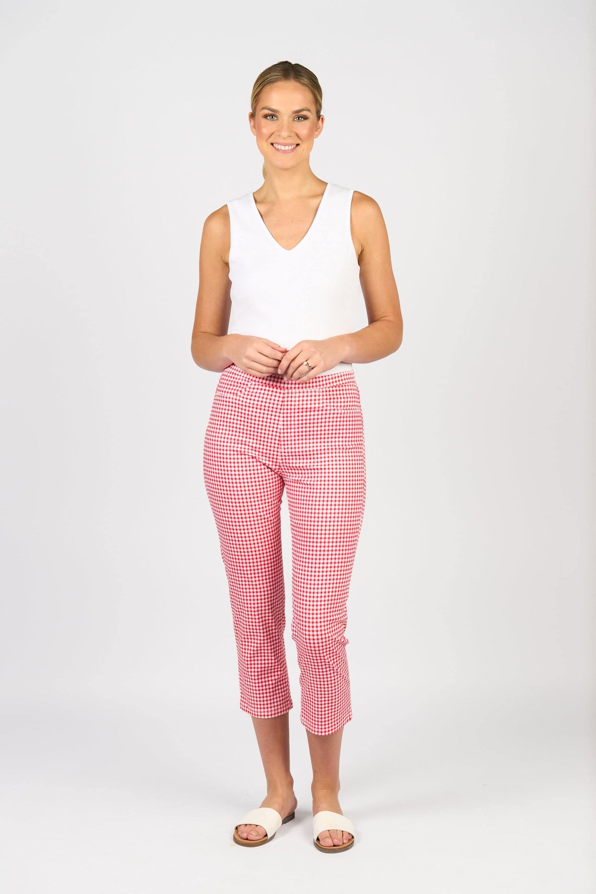 A woman smiles against a plain background, wearing a sleeveless white top, Sierra 7/8 Pant in Red Gingham by Vassalli, and white slide sandals.