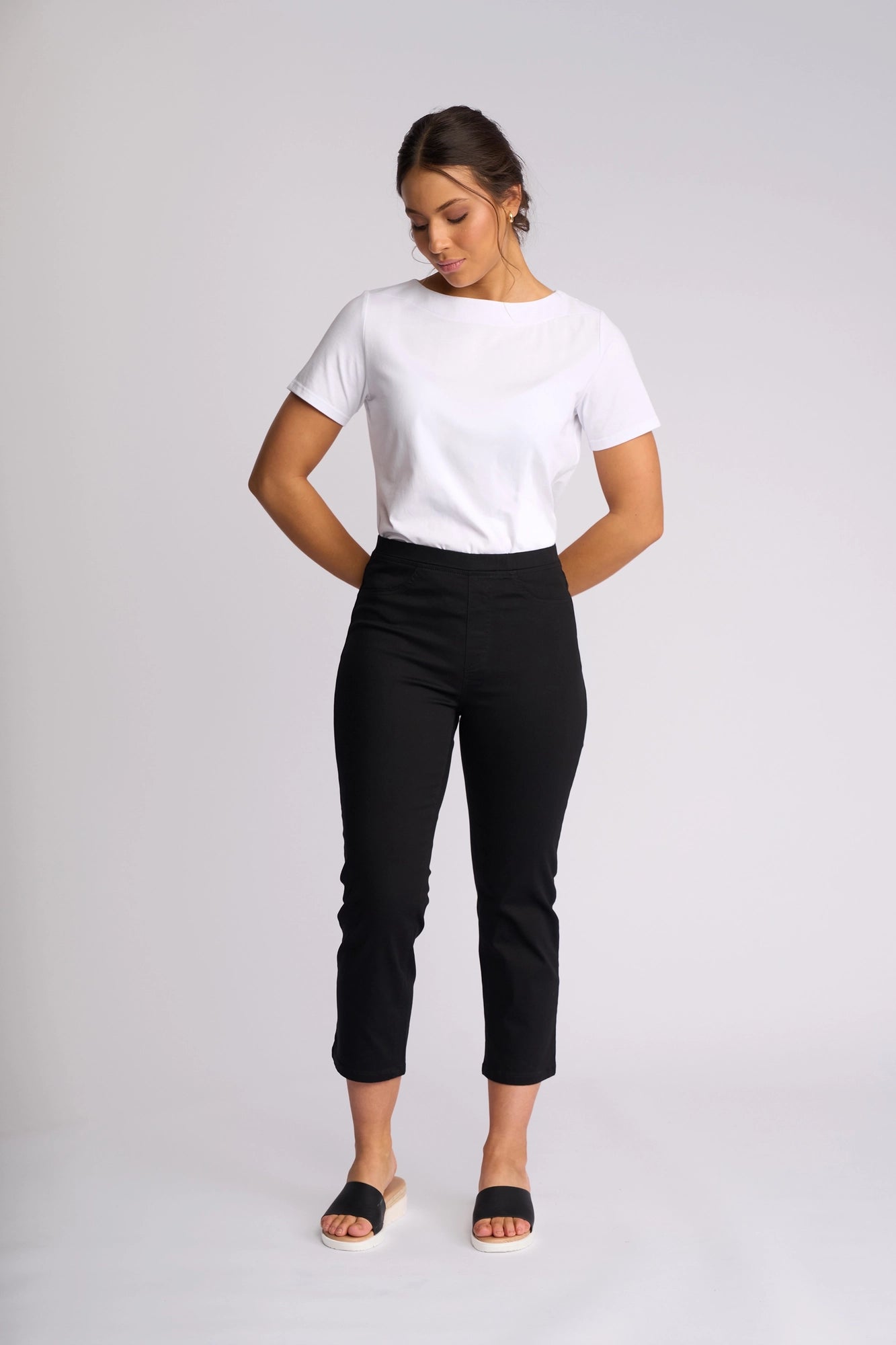 A woman stands against a plain background, wearing a white t-shirt and the Vassalli Sierra 7/8 Pant in Black—sleek, slim-leg pull-on pants crafted from stretch cotton—paired with black slide sandals. Her hands are behind her back, and she gazes downward with a neutral expression.