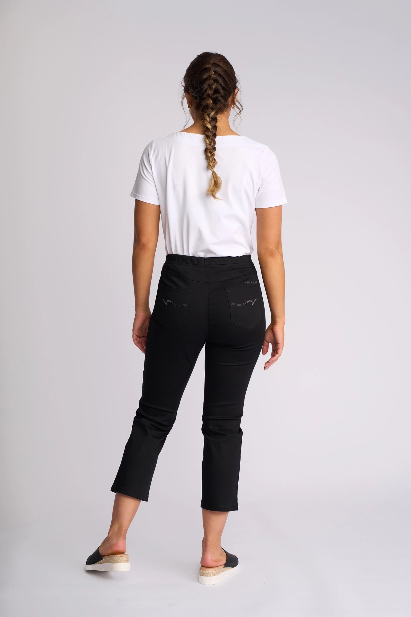 A woman with braided hair stands facing away, wearing a white t-shirt and the Vassalli Sierra 7/8 Pant in black—a comfortable pull-on slim-leg style—paired with black slip-on shoes, against a plain light background.