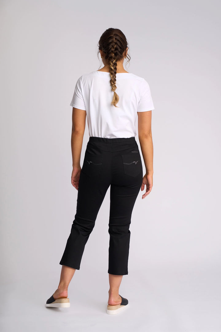 A woman with braided hair stands facing away, wearing a white t-shirt and the Vassalli Sierra 7/8 Pant in black—a comfortable pull-on slim-leg style—paired with black slip-on shoes, against a plain light background.