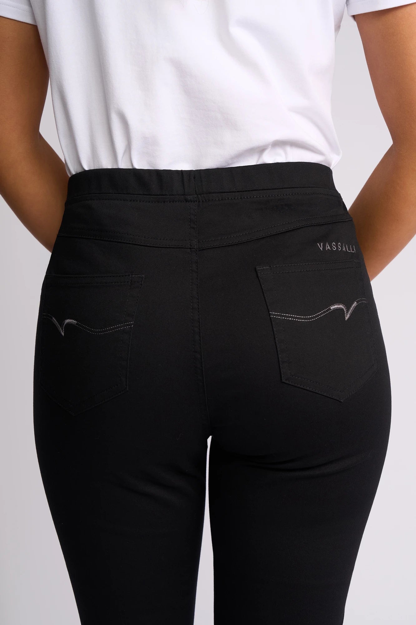 A person wearing a white shirt and the Vassalli Sierra 7/8 Pant in Black is shown from the back, with emphasis on the waist and hip area. The slim-leg pant, made from comfortable stretch cotton, features two back pockets with decorative stitching and is subtly finished with a small VASSALLI logo.