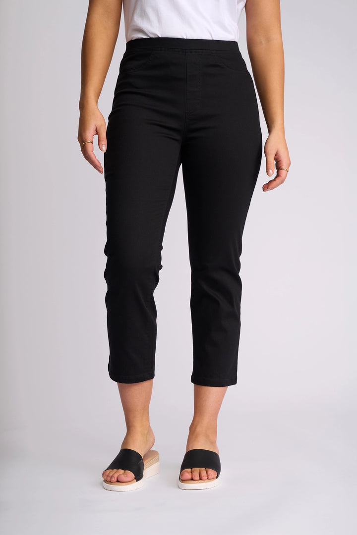 A person wearing a white shirt, Vassalli’s Sierra 7/8 Pant in black—a slim-leg, stretch cotton, pull-on style—and black slide sandals with a light platform stands against a plain light background.