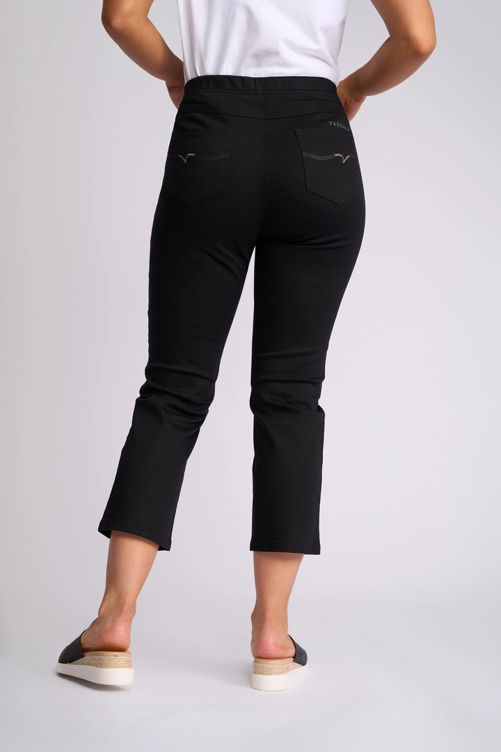 A person stands facing away, wearing the Vassalli Sierra 7/8 Pant in black, paired with a white shirt and black slip-on shoes with light soles, against a plain light background.