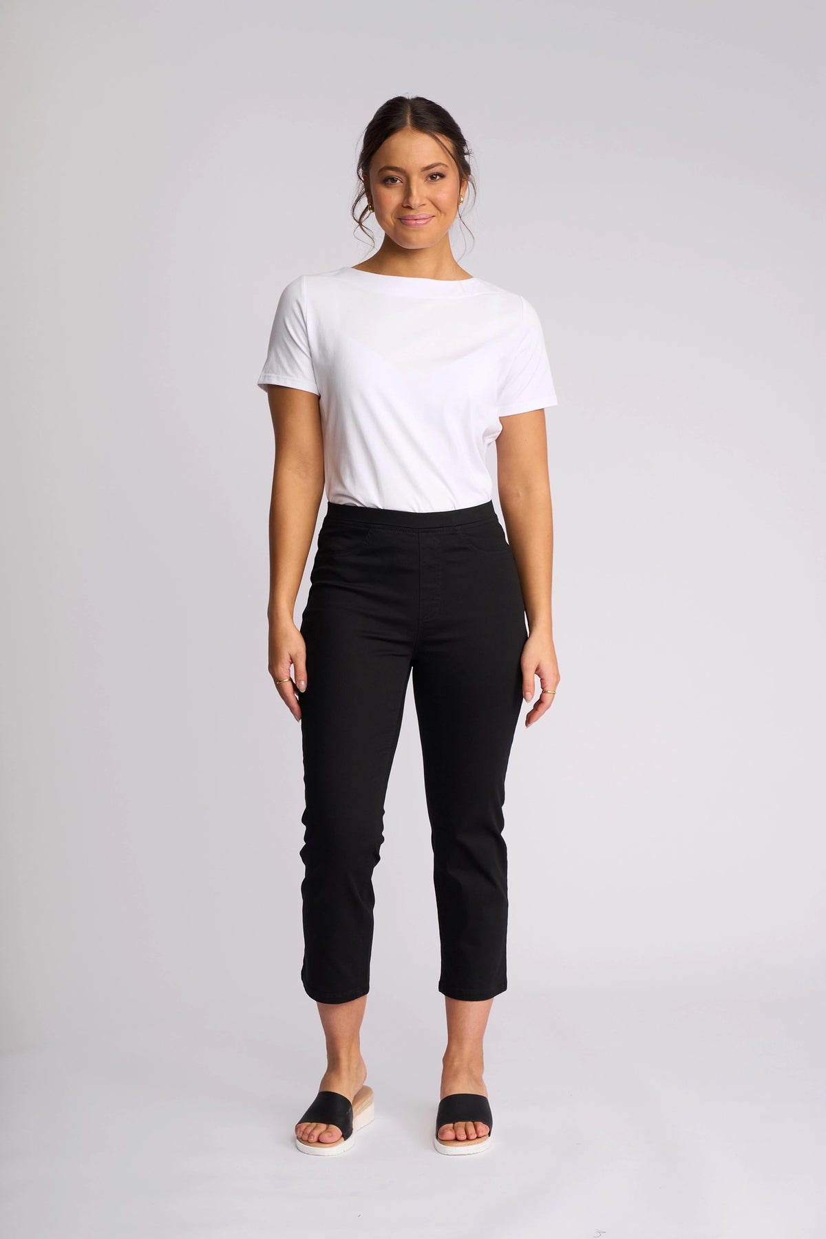 A person stands against a plain white background, wearing a white t-shirt paired with Vassalli’s Sierra 7/8 Pant in Black—stretch cotton slim-leg pants featuring an easy pull-on style—along with black slide sandals. Their hair is tied back and they have a relaxed expression.