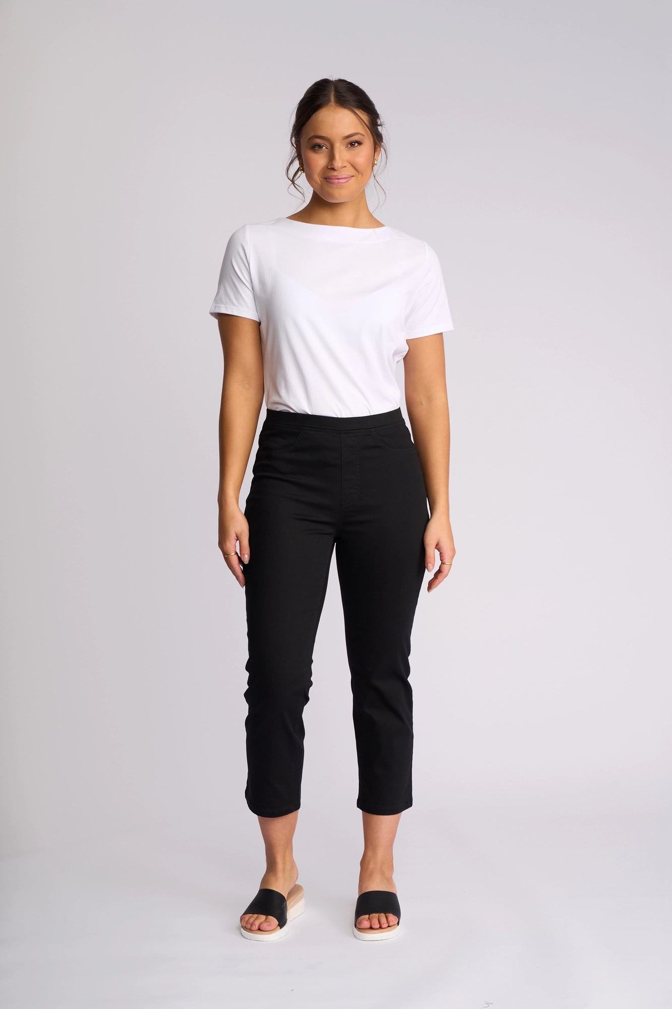 A person stands against a plain white background, wearing a white t-shirt paired with Vassalli’s Sierra 7/8 Pant in Black—stretch cotton slim-leg pants featuring an easy pull-on style—along with black slide sandals. Their hair is tied back and they have a relaxed expression.