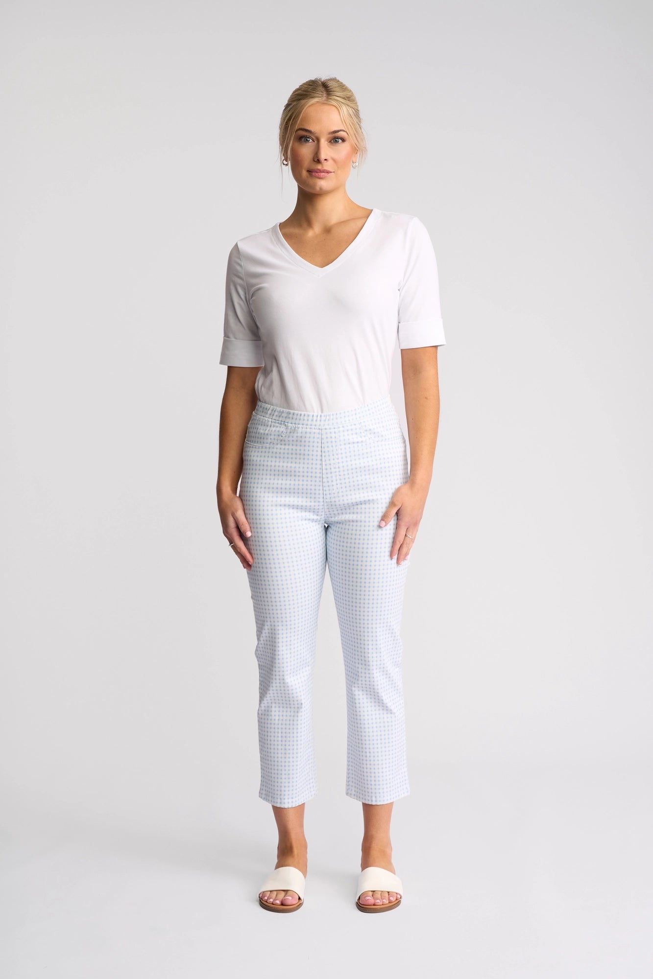 A woman stands facing forward against a plain white background, wearing a white V-neck top and the Vassalli Sierra 7/8 Pant in Blue Gingham, crafted from stretch cotton in a pull-on slim leg style, paired with white slide sandals.