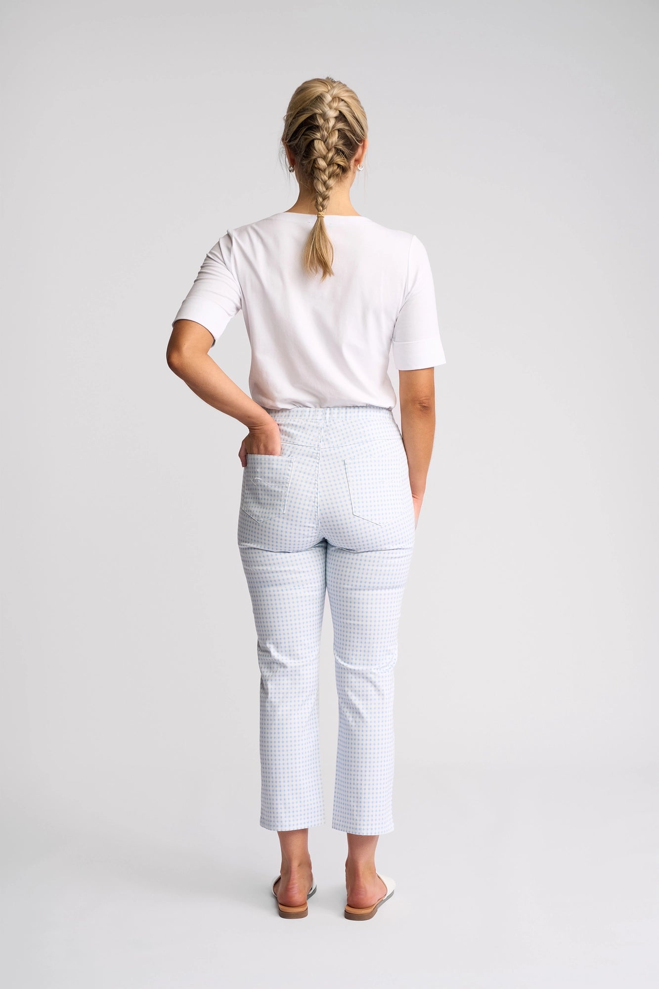 A woman with blonde hair in a braid stands facing away, wearing a white short-sleeve shirt, white sandals, and the Vassalli Sierra 7/8 Pant in Blue Gingham—pull-on slim leg pants crafted from stretch cotton—against a plain white background.
