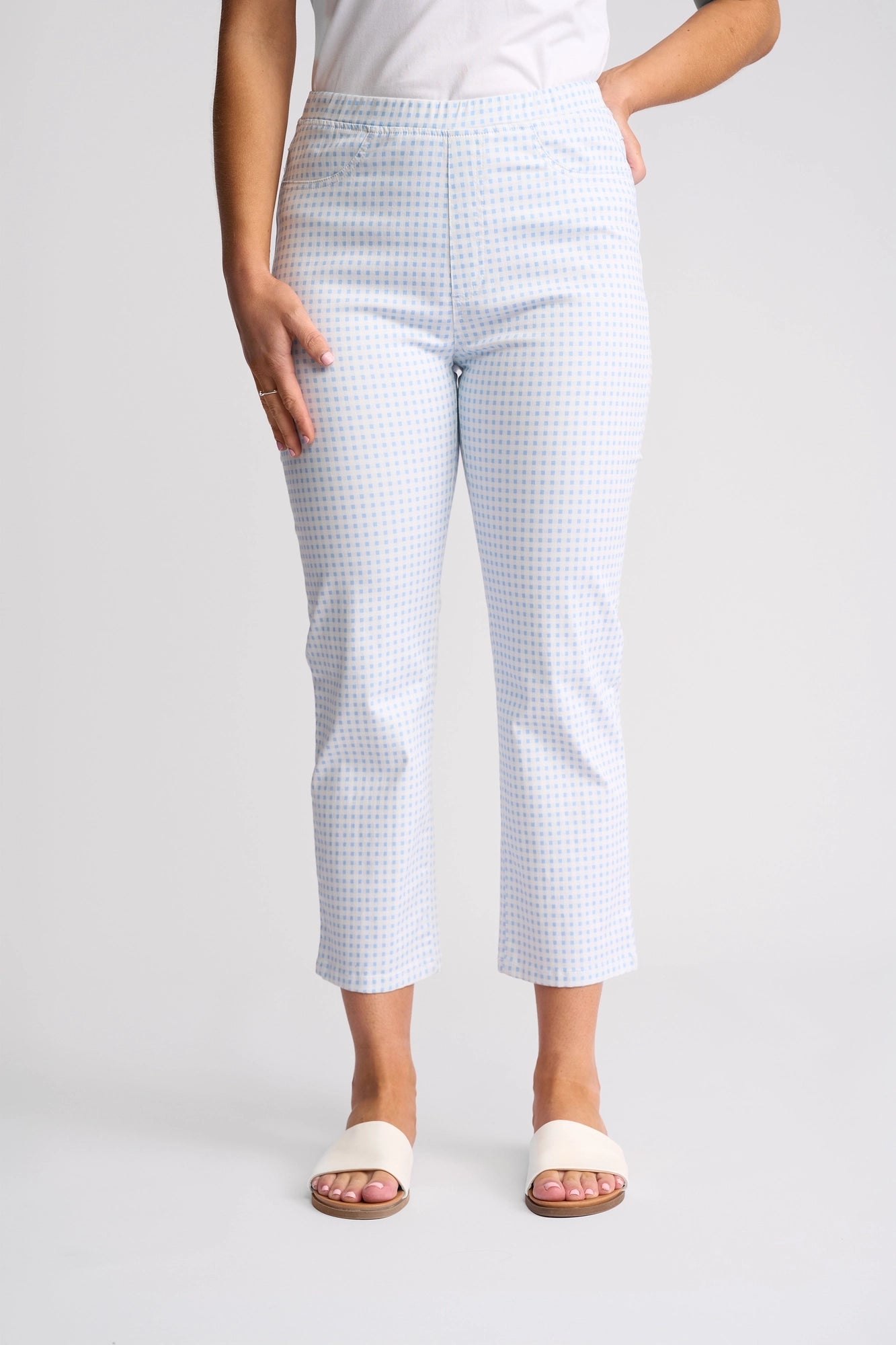 A person wearing the Vassalli Sierra 7/8 Pant in Blue Gingham—slim leg, ankle-length pull-on pants crafted from stretch cotton—paired with a white top and white open-toe sandals, stands against a plain light background.